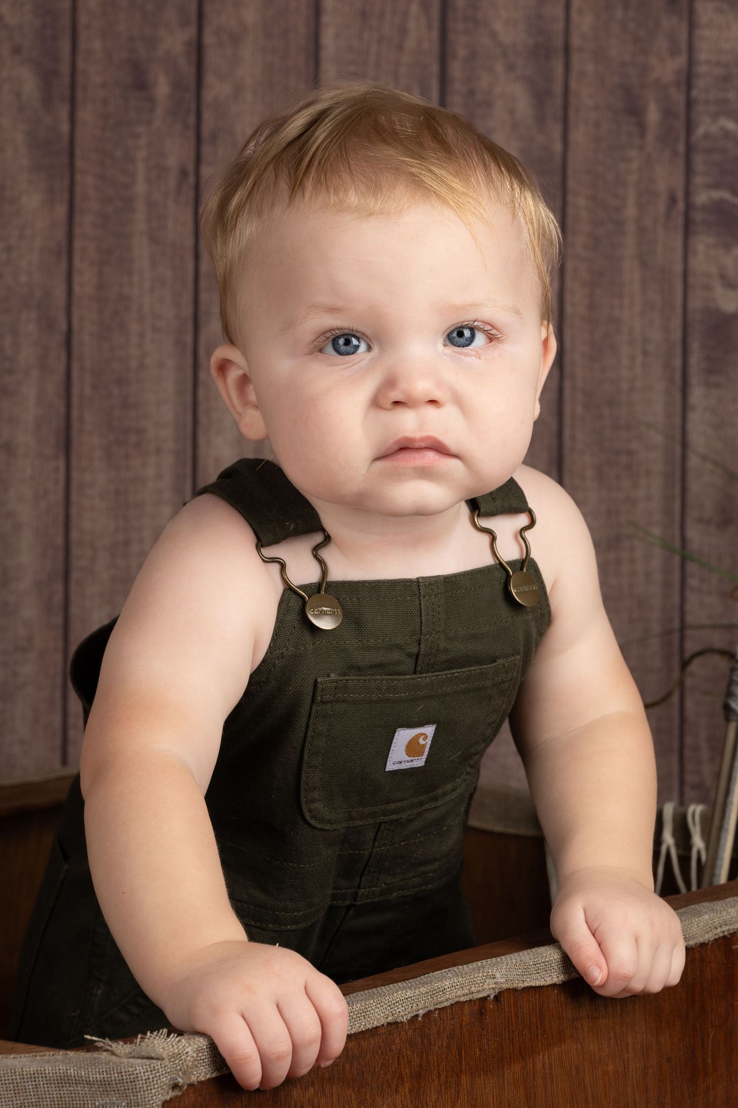 Adorable baby with blue eyes wearing olive green Carhartt overalls, posed in a rustic wooden crate setting.