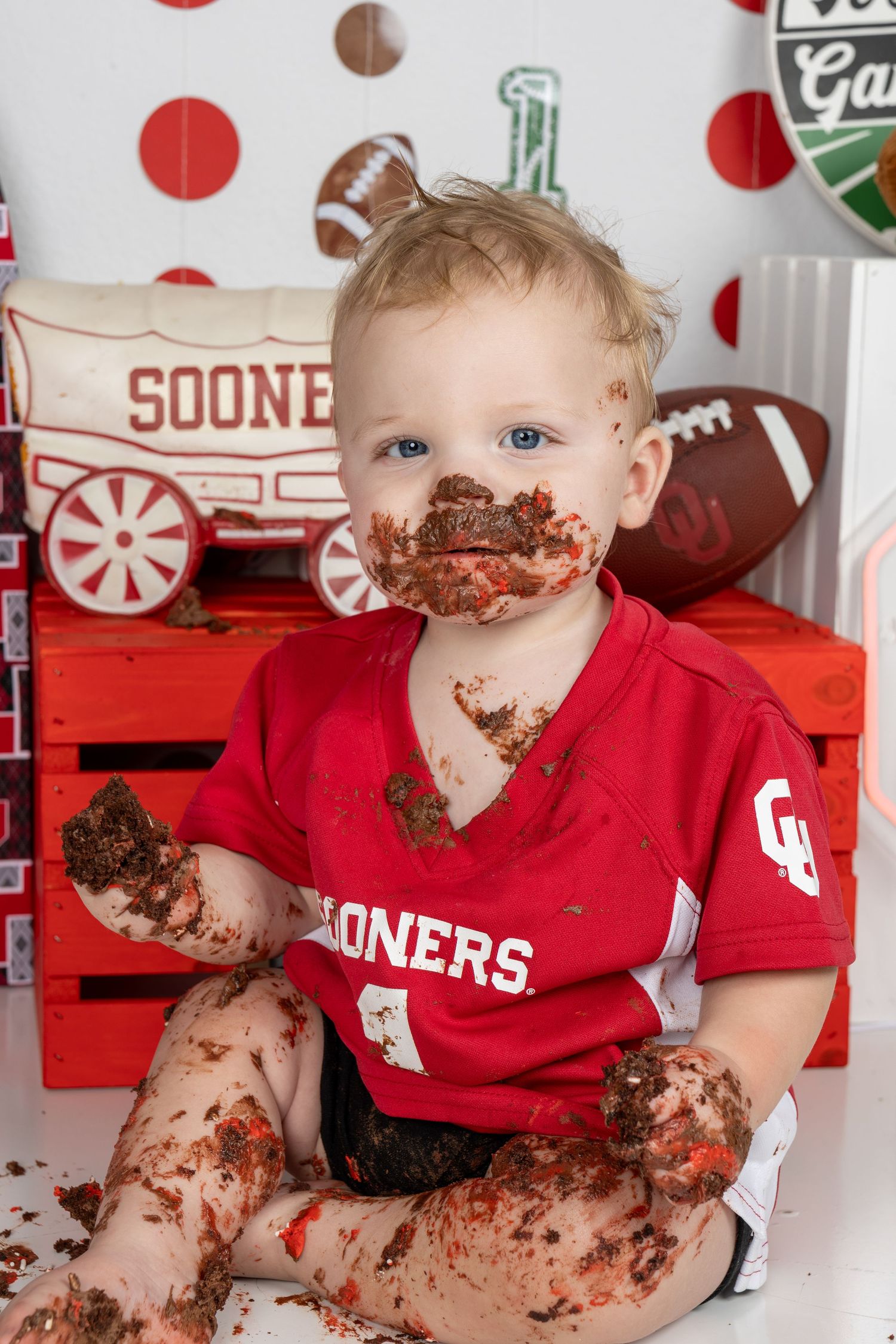 Baby boy in Oklahoma Sooners jersey covered in chocolate cake during first birthday smash cake photo session.