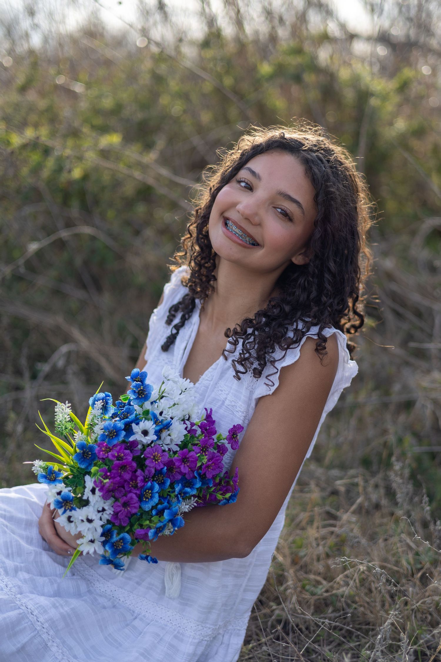 A person in a white dress poses with a vibrant blue and purple bouquet in an outdoor natural setting.