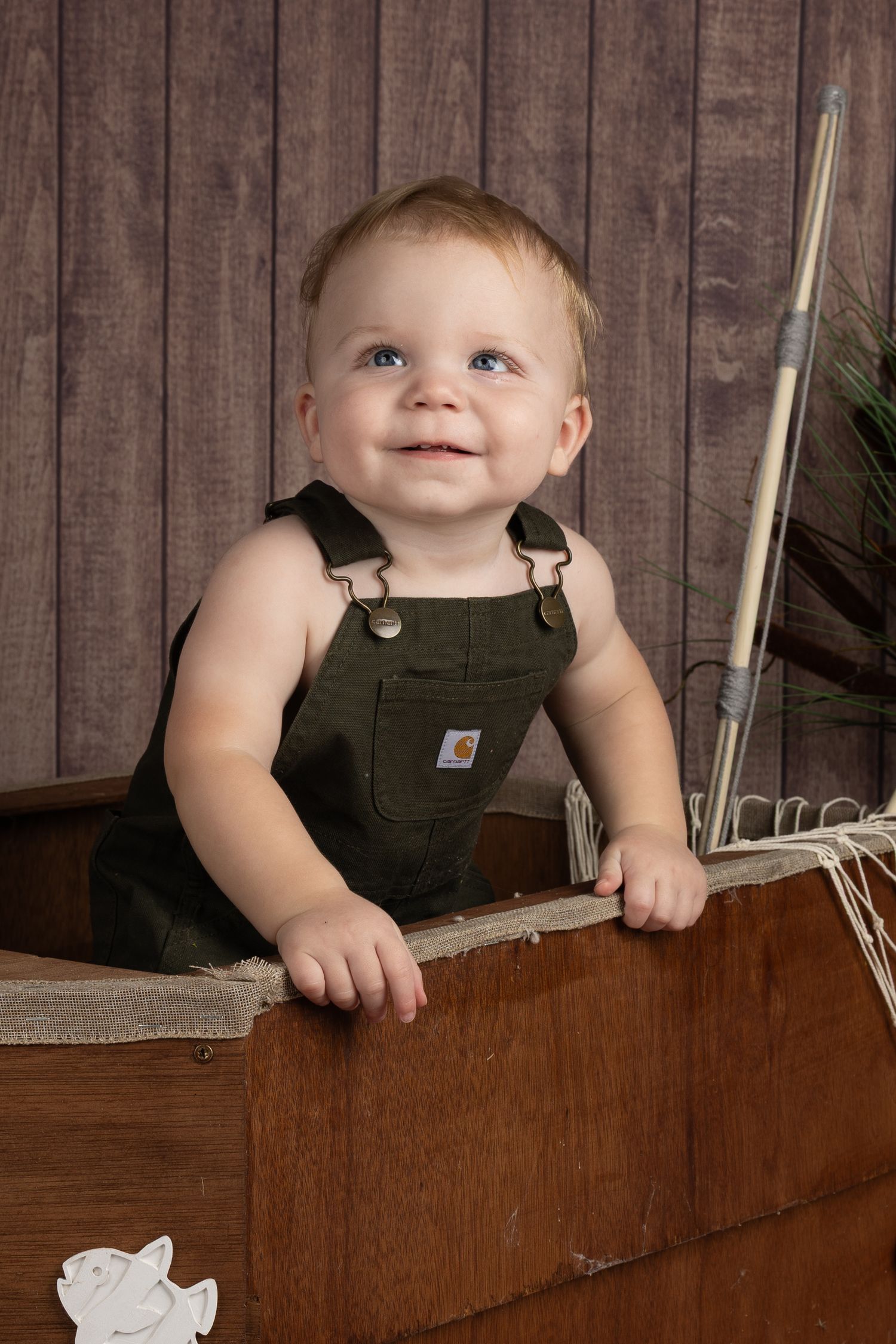 Smiling baby in green Carhartt overalls sitting in a rustic wooden boat with a fishing pole, posed against a wood backdrop.
