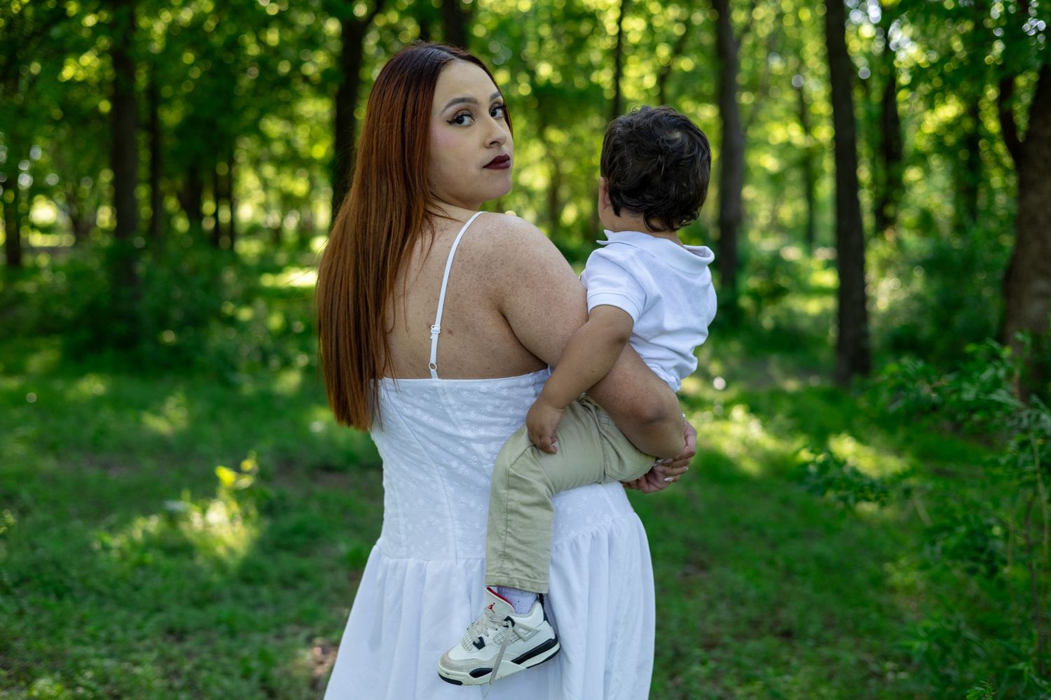 A young woman in a white dress holds a toddler boy in a lush green forest setting.