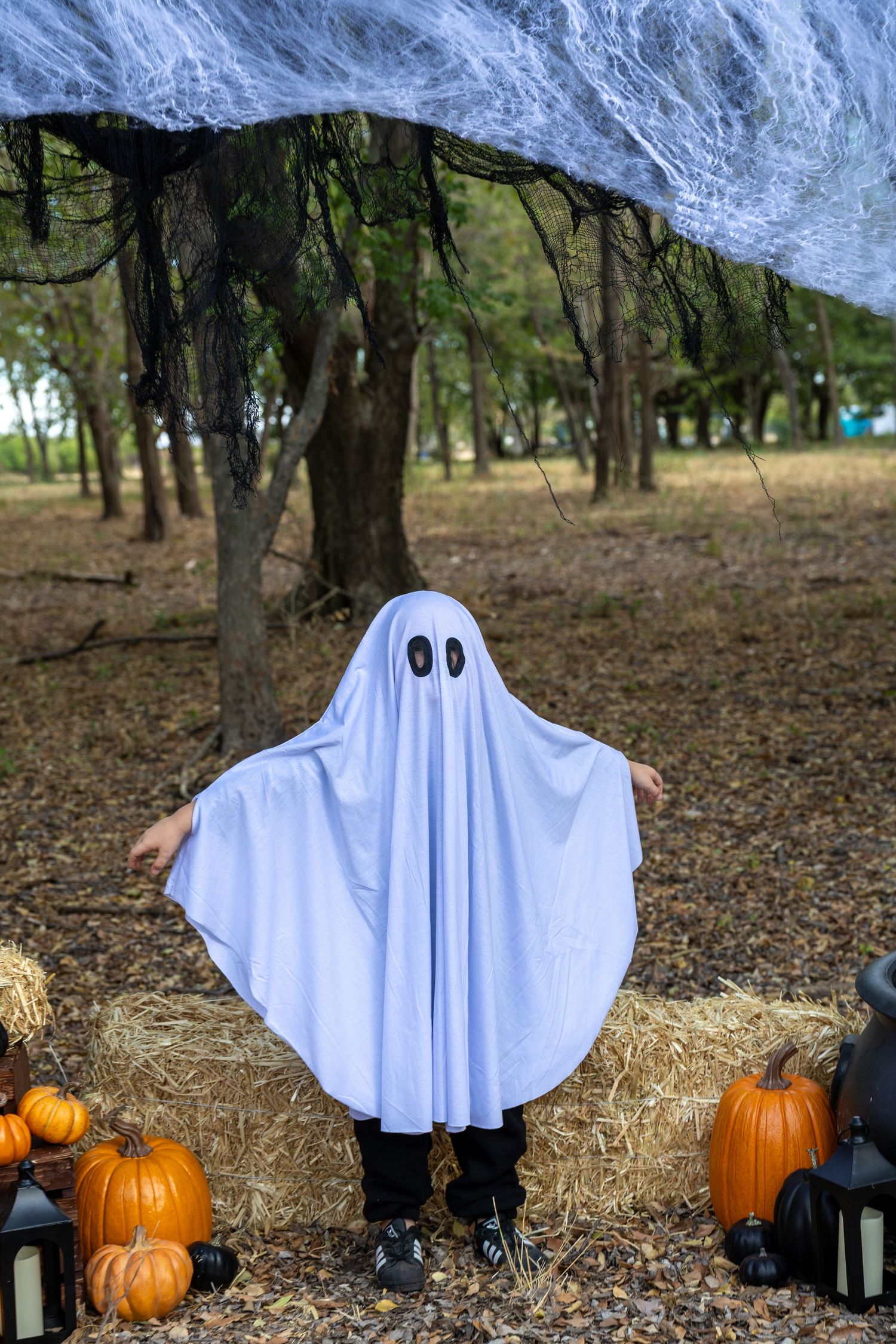 Child in white ghost costume with black eyes stands among pumpkins and hay bales in a Halloween forest setting.