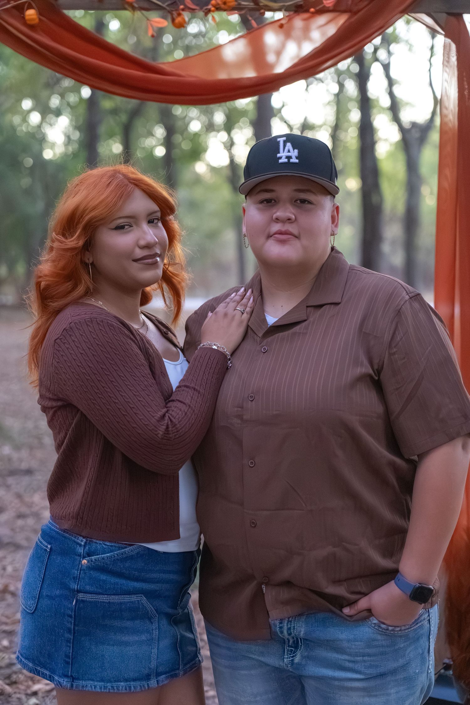 Couple posing outdoors under orange floral arch, woman with red hair in brown cardigan, man in LA cap and brown shirt.