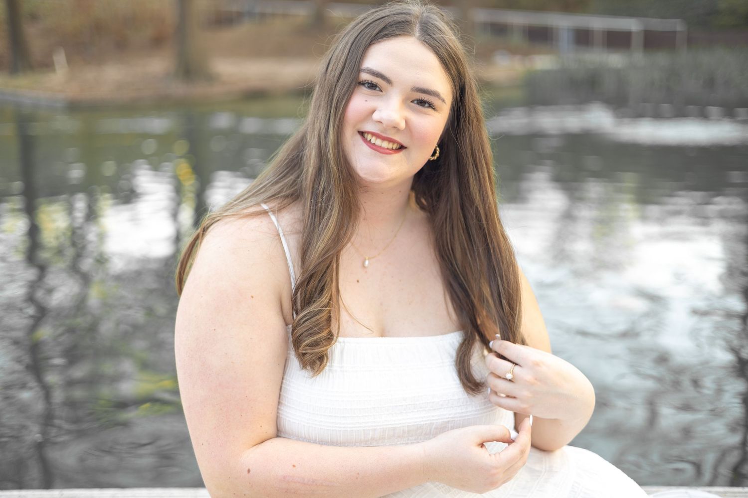 A smiling person in a white top poses outdoors by a tranquil pond on a sunny day.