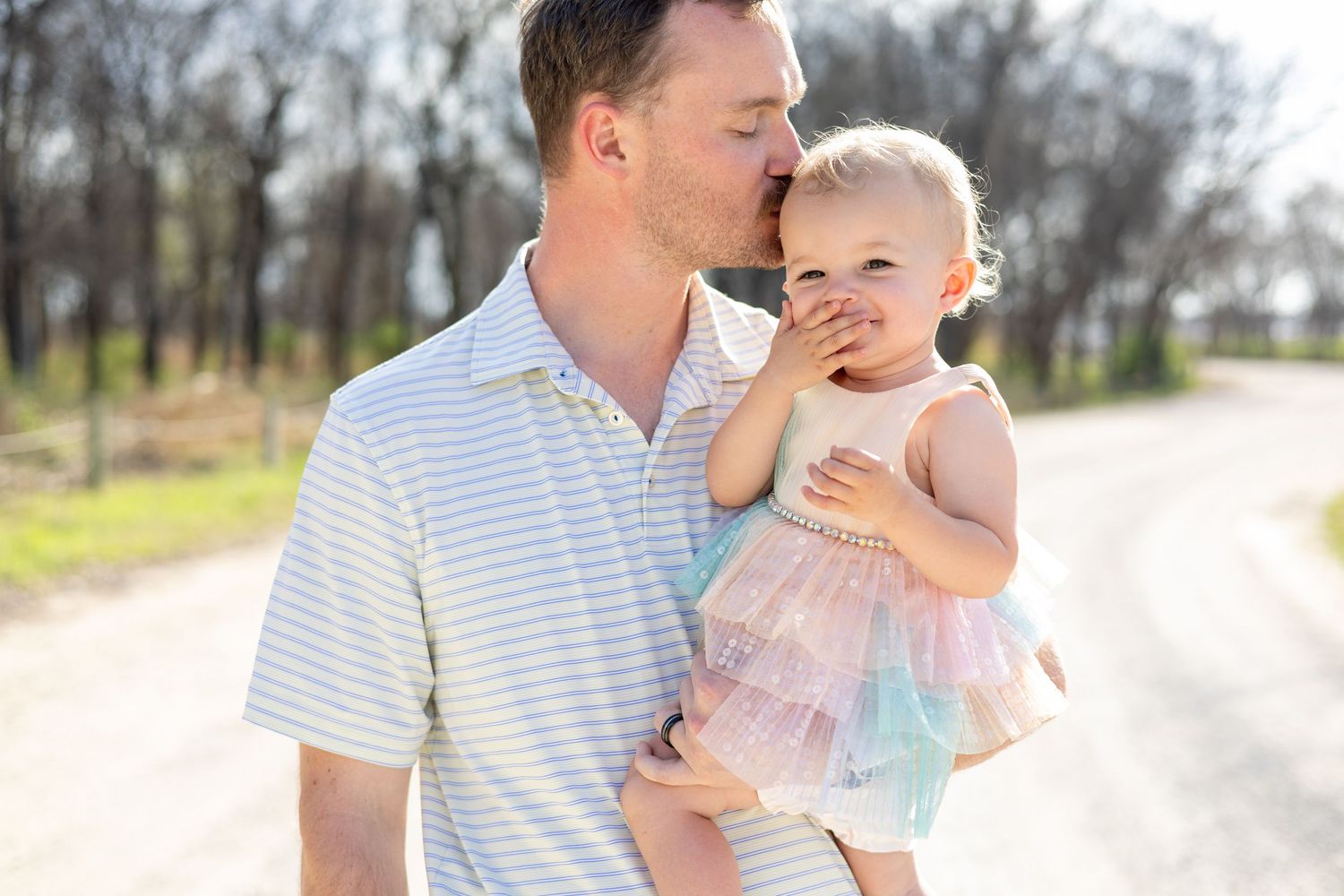 A parent and baby in matching striped shirts share tender moments outdoors during a sunny photoshoot.