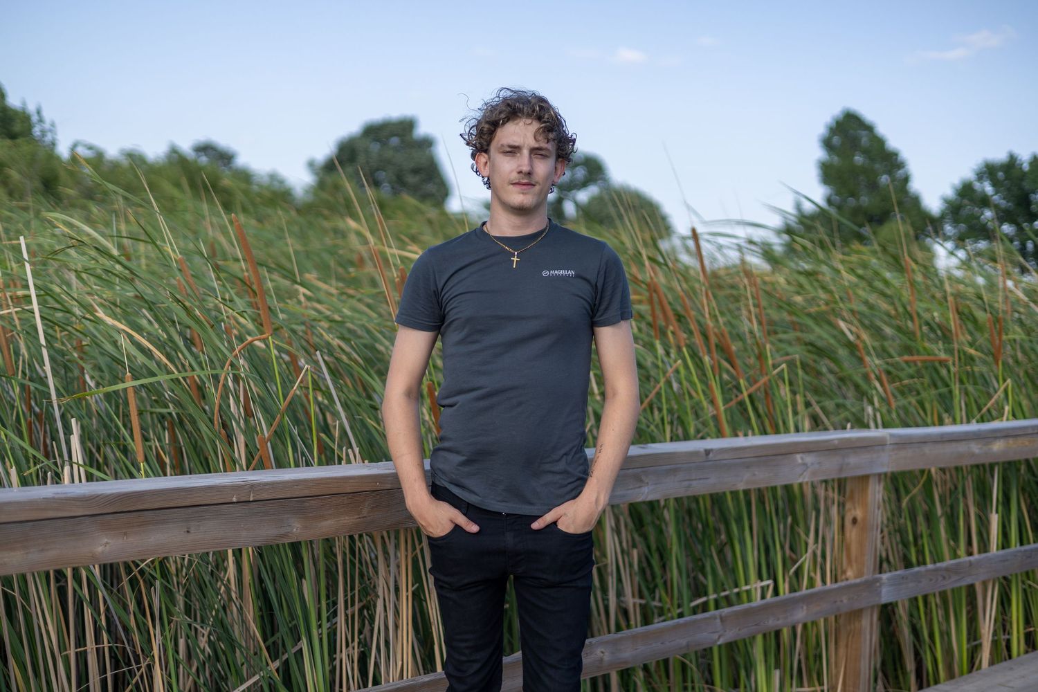 Young man in gray t-shirt standing on wooden bridge surrounded by tall reeds and green trees at dusk.