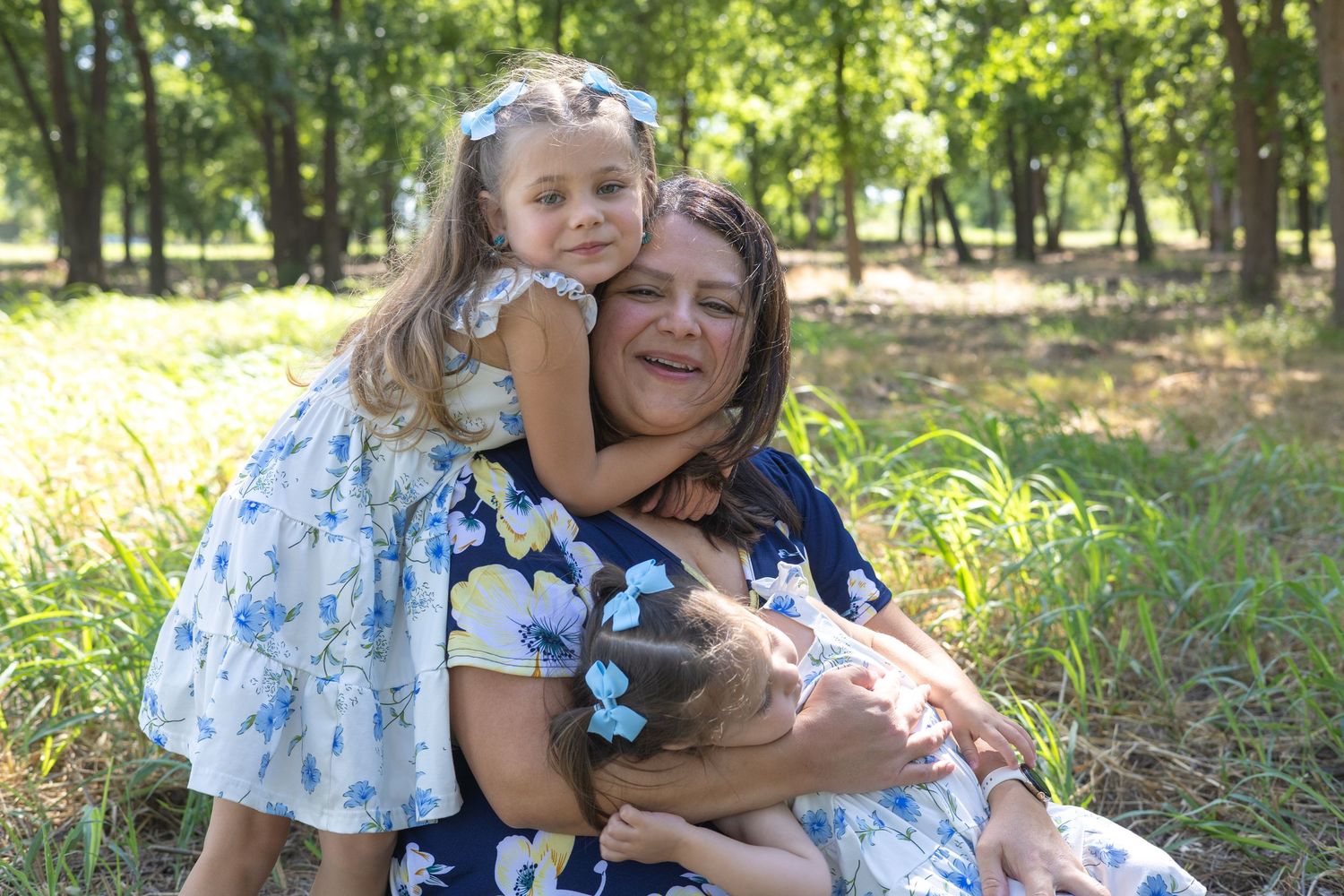 A woman sits outdoors hugging two young girls in floral dresses, smiling joyfully in a lush green park setting.
