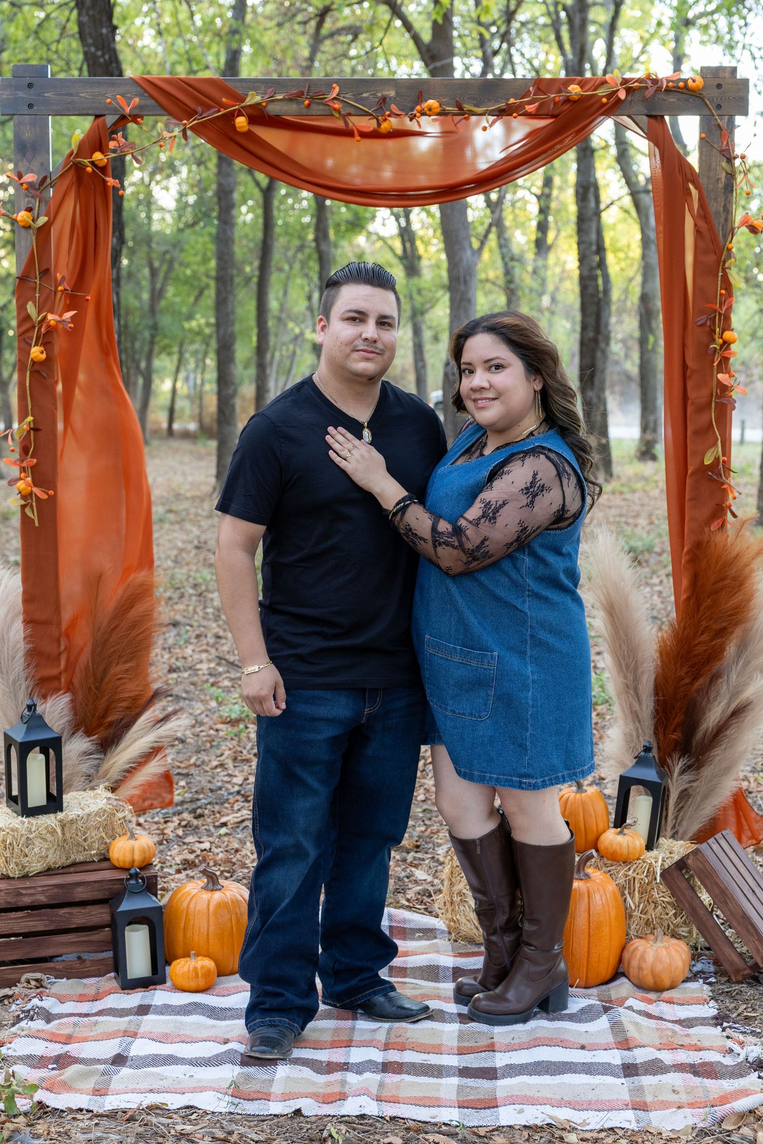 Couple poses under rustic fall arch with pumpkins, hay bales, and orange draping in an autumn outdoor setting.