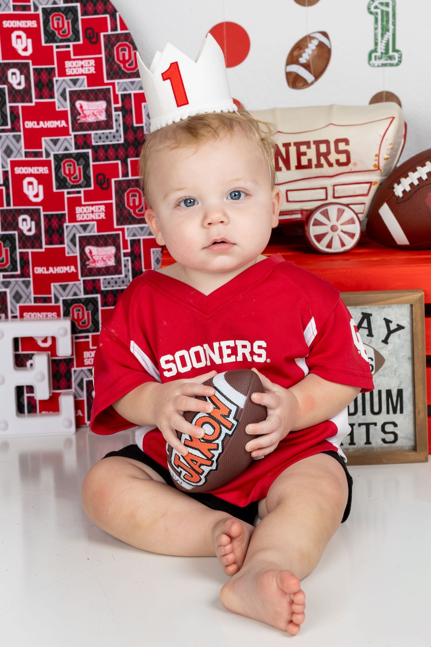 Baby in Sooners jersey holds football, wearing birthday crown, surrounded by Oklahoma Sooners themed decorations.