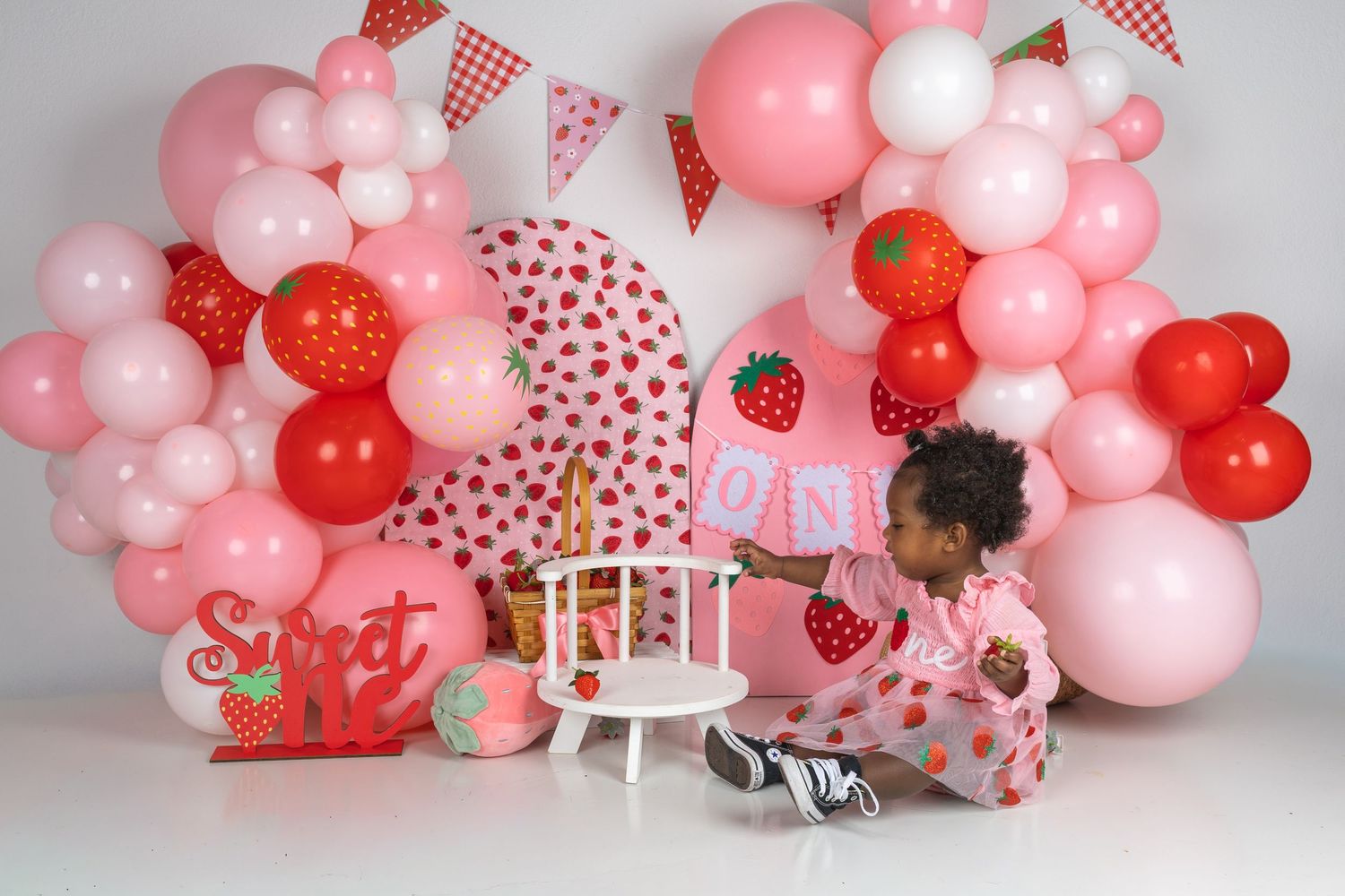 Toddler girl at a strawberry-themed first birthday party with pink and red balloons and Sweet One decorations.