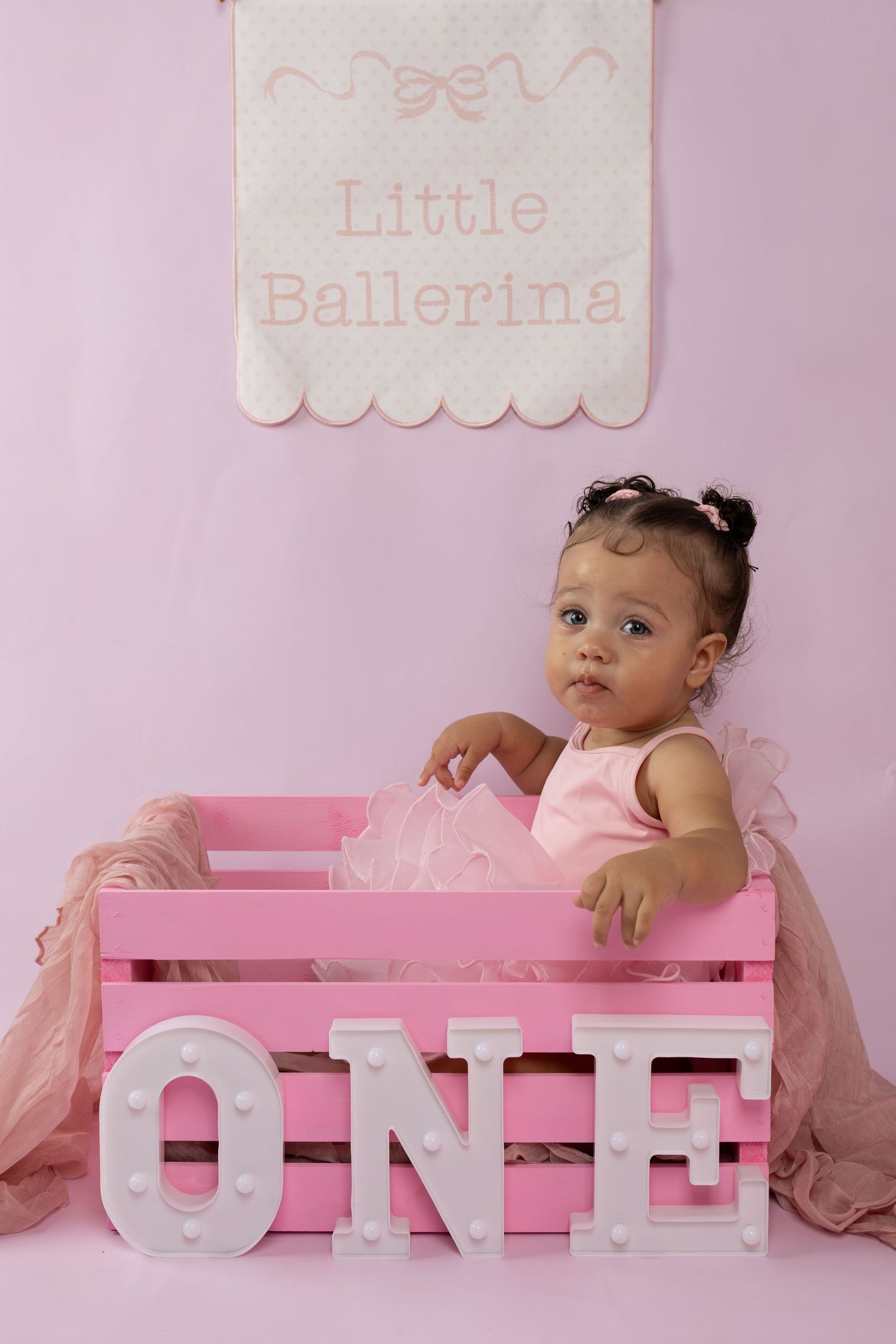 Adorable baby girl in pink tutu sits in pink crate with ONE marquee letters for first birthday photo.