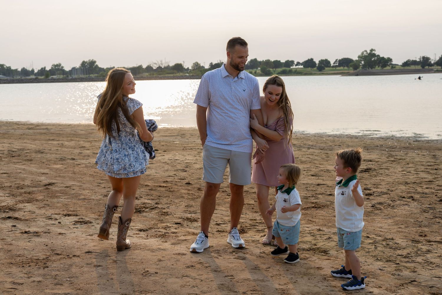 Family of five enjoying a sunny beach walk near calm water, with two young children and parents dressed in casual summer outfits.