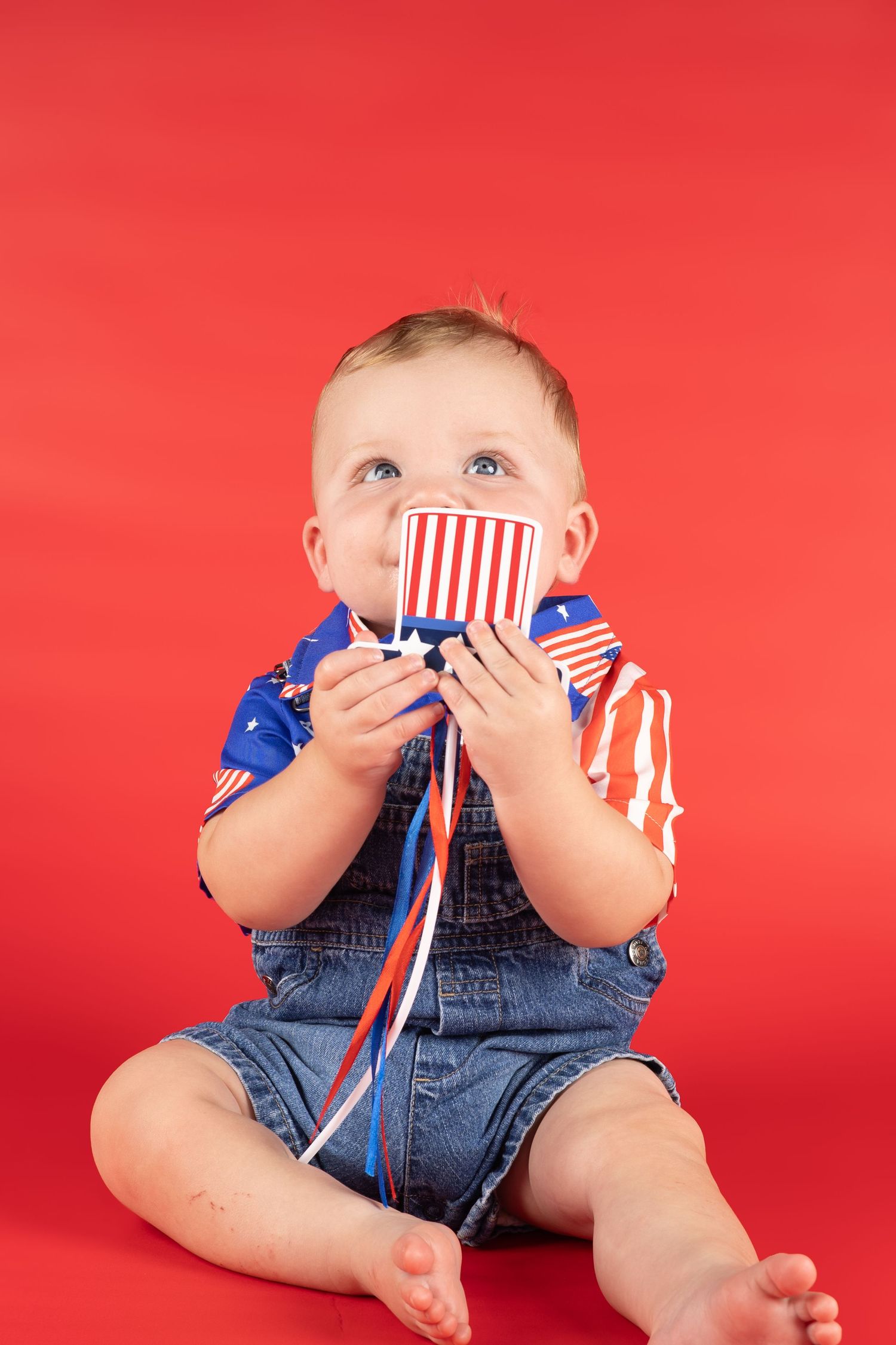 Baby in denim overalls and patriotic shirt holds red white blue cup against red background.