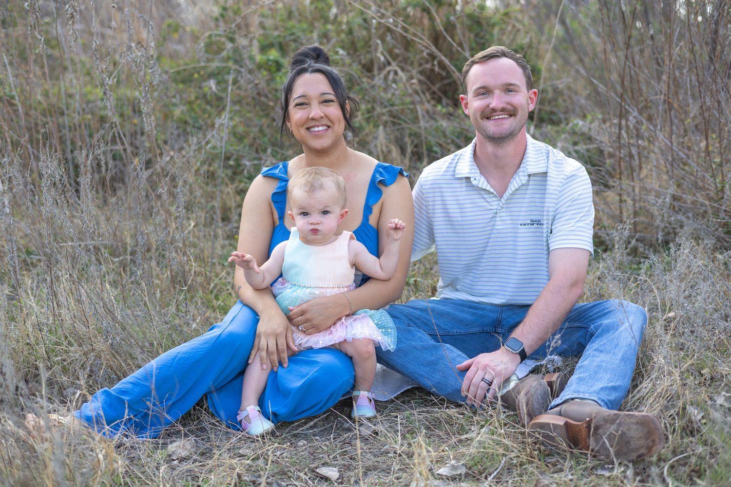 A family relaxes together on a grassy hillside wearing coordinated blue and white outfits for an outdoor photo session.