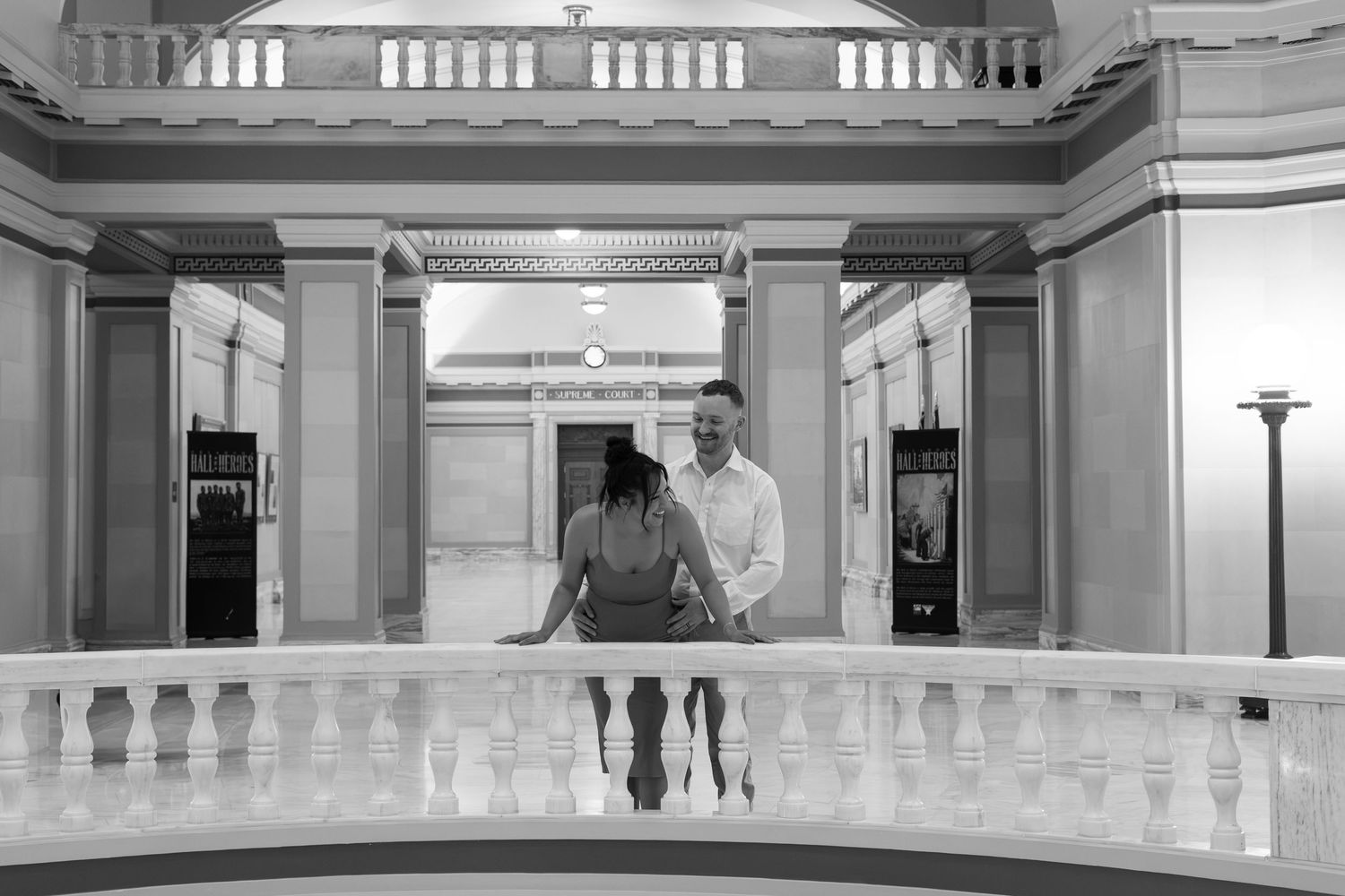 Couple smiling together on elegant balcony with white balustrade and grand columns in black and white photo.