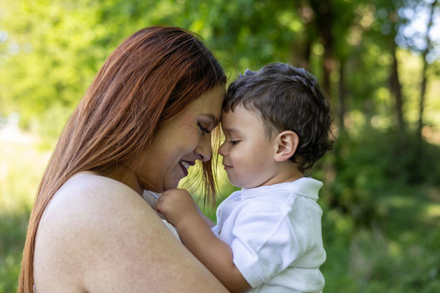 A mother with red hair tenderly holds her toddler, touching foreheads in a loving moment outdoors.