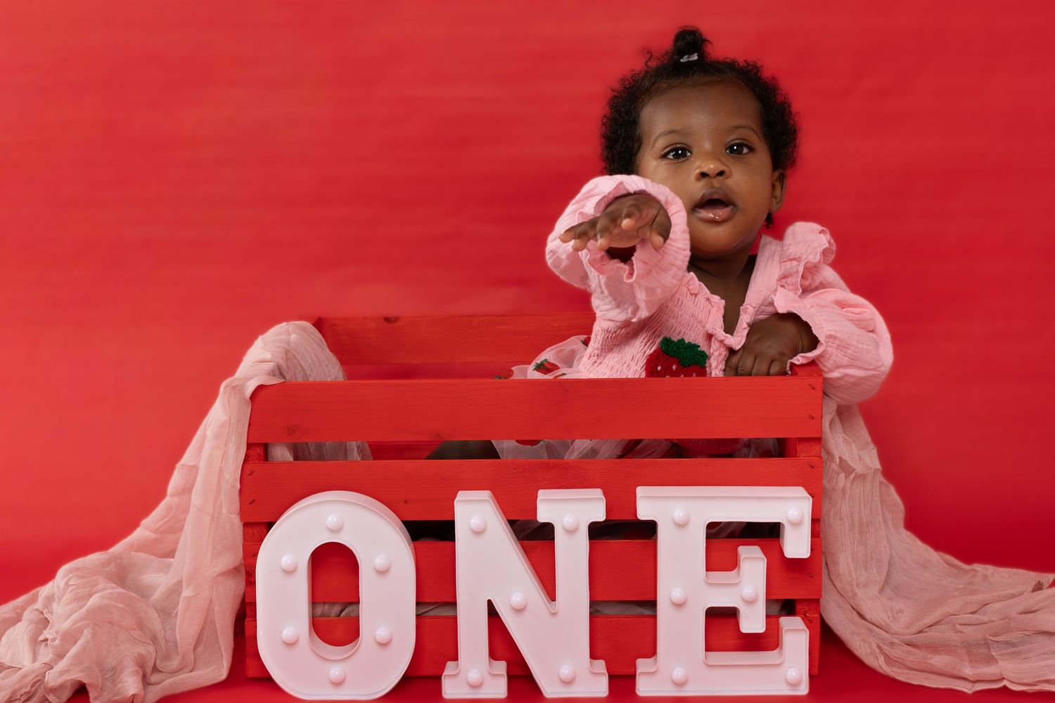 Adorable baby girl in pink outfit sitting in red crate with marquee ONE letters for first birthday photo.