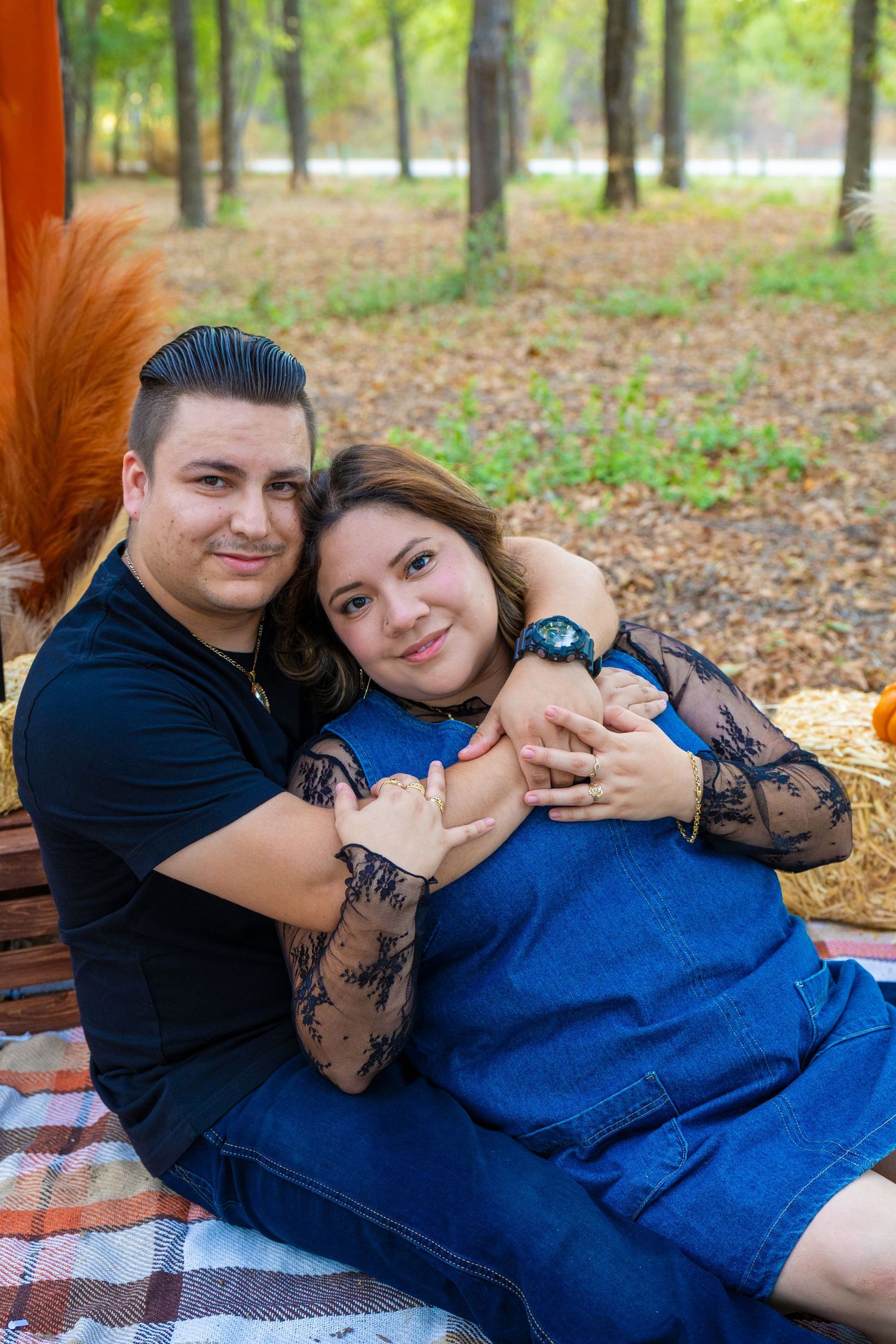 A couple shares a warm embrace on a plaid blanket during an autumn picnic in a park with fall foliage.