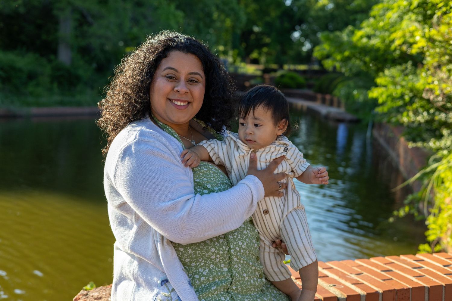 A smiling woman with curly hair holds a baby outdoors near a pond surrounded by lush green trees.