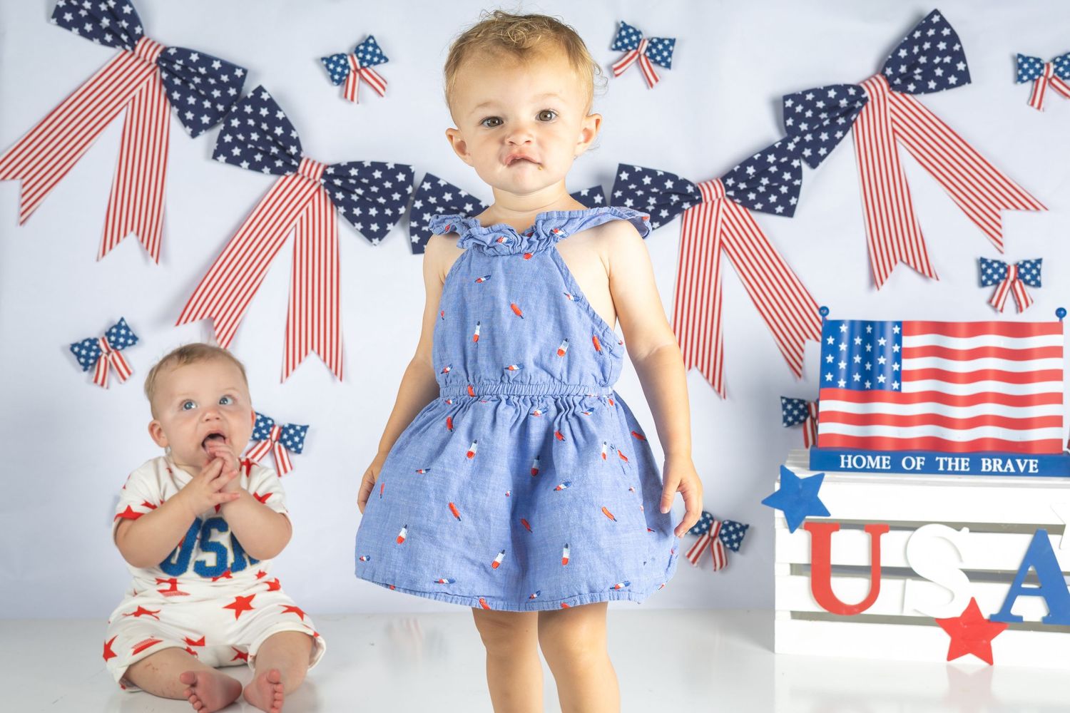 Two toddlers pose in patriotic outfits with American flag decorations and USA sign for Fourth of July photos.