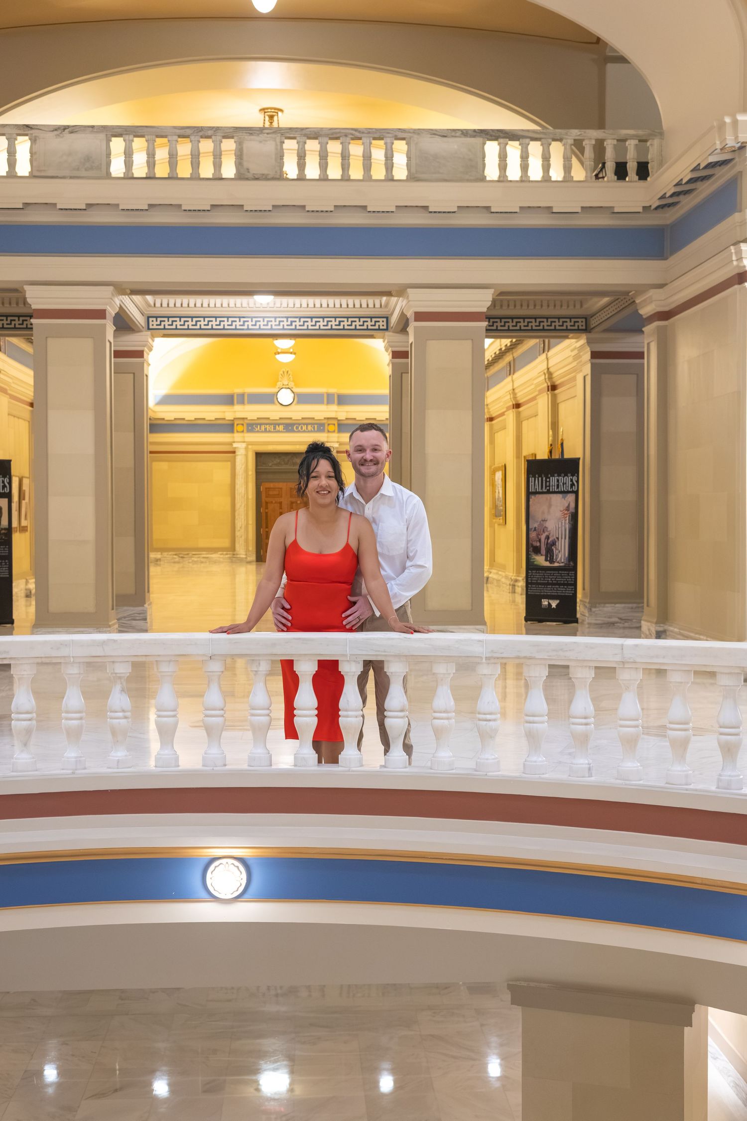 Couple posing on ornate white balcony in elegant classical building interior with arched ceilings.