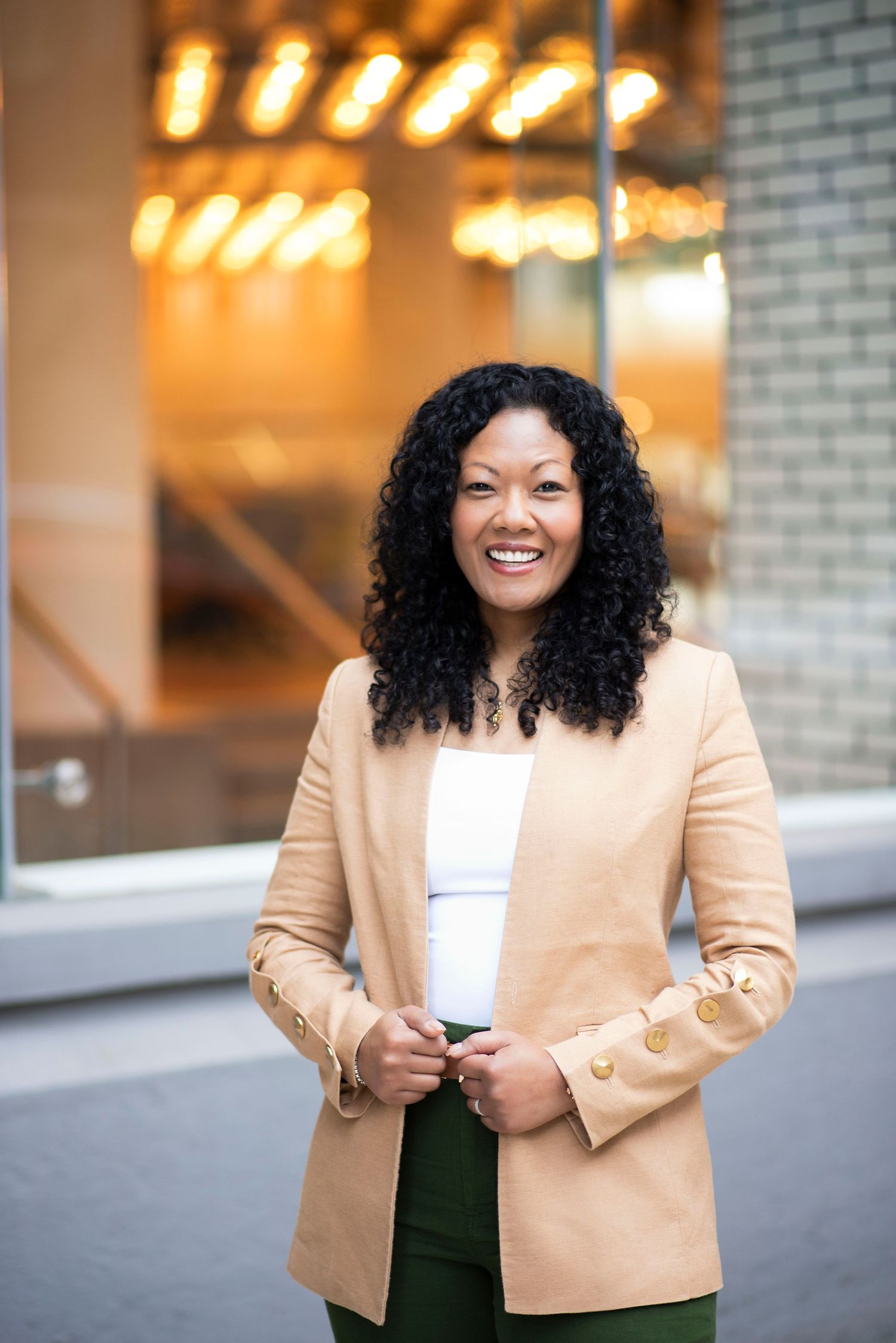 Professional wearing tan blazer and dark top poses confidently in front of illuminated building.