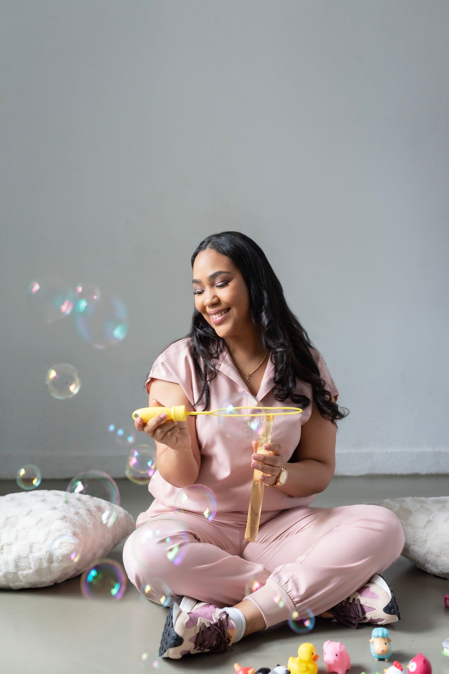 Playful portrait session with bubbles and toys in pink loungewear against gray background.