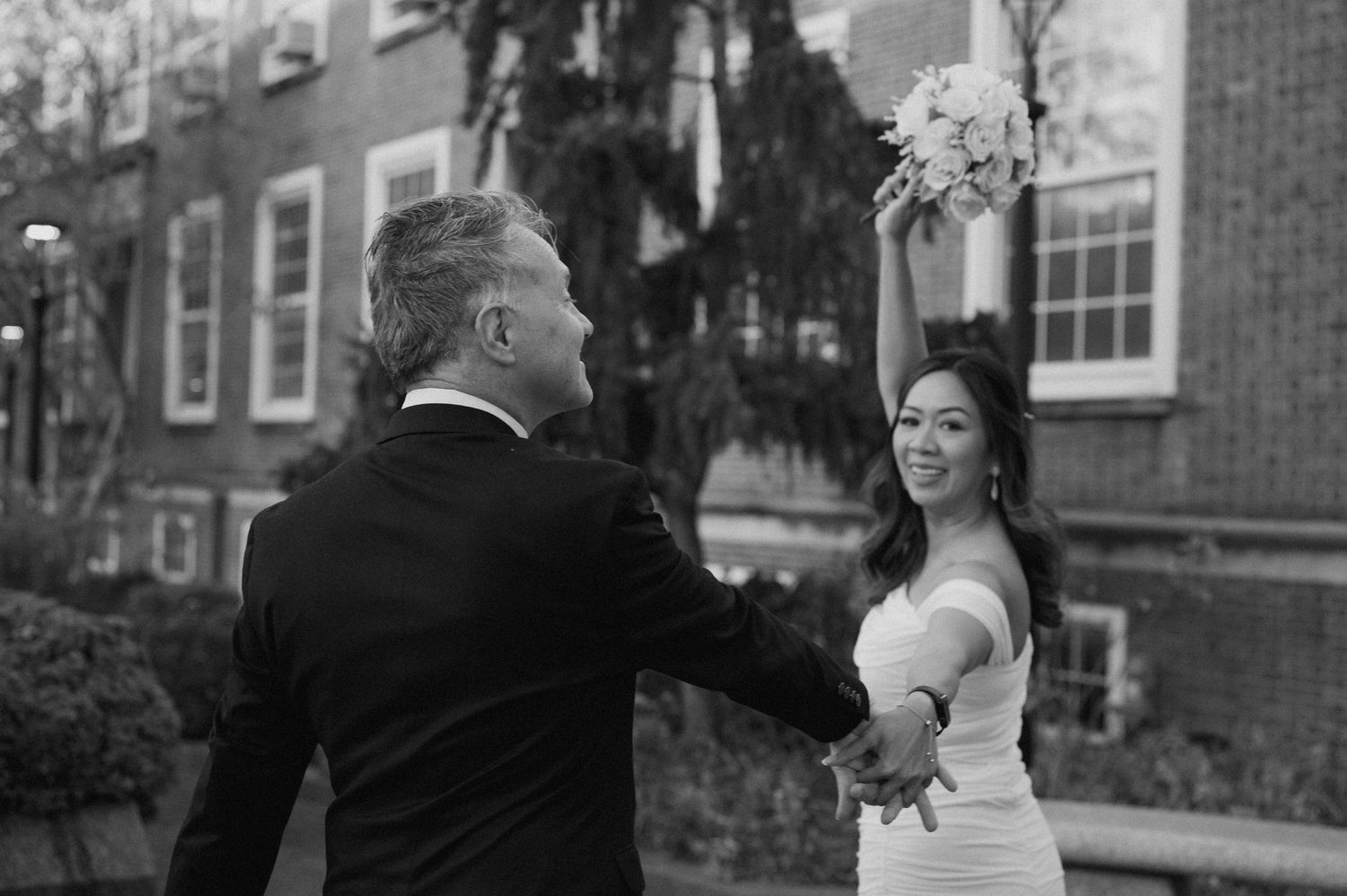 A romantic outdoor wedding scene with brick buildings and trees in the background.