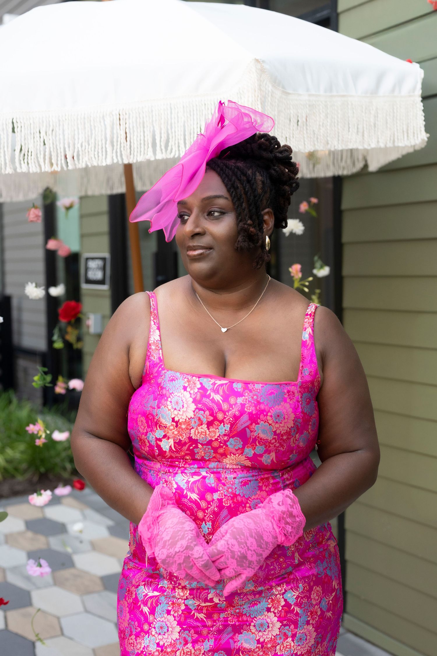 Woman in pink floral dress and fascinator hat poses outdoors under white fringed umbrella.