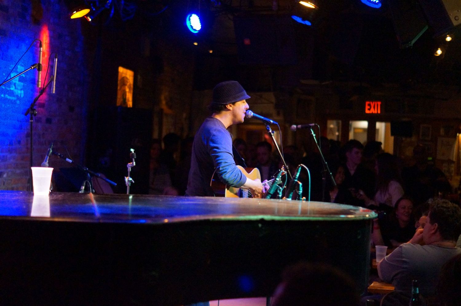 Musician performs at dimly lit venue, playing keyboard at microphone stand with audience seated in background.