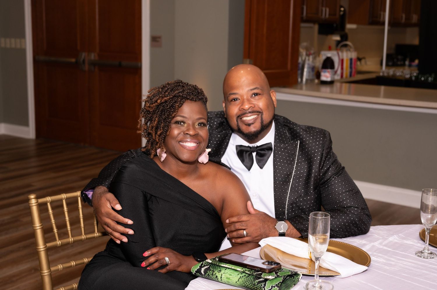 Couple in formal black attire smiling at a banquet table with champagne glasses at an elegant event.