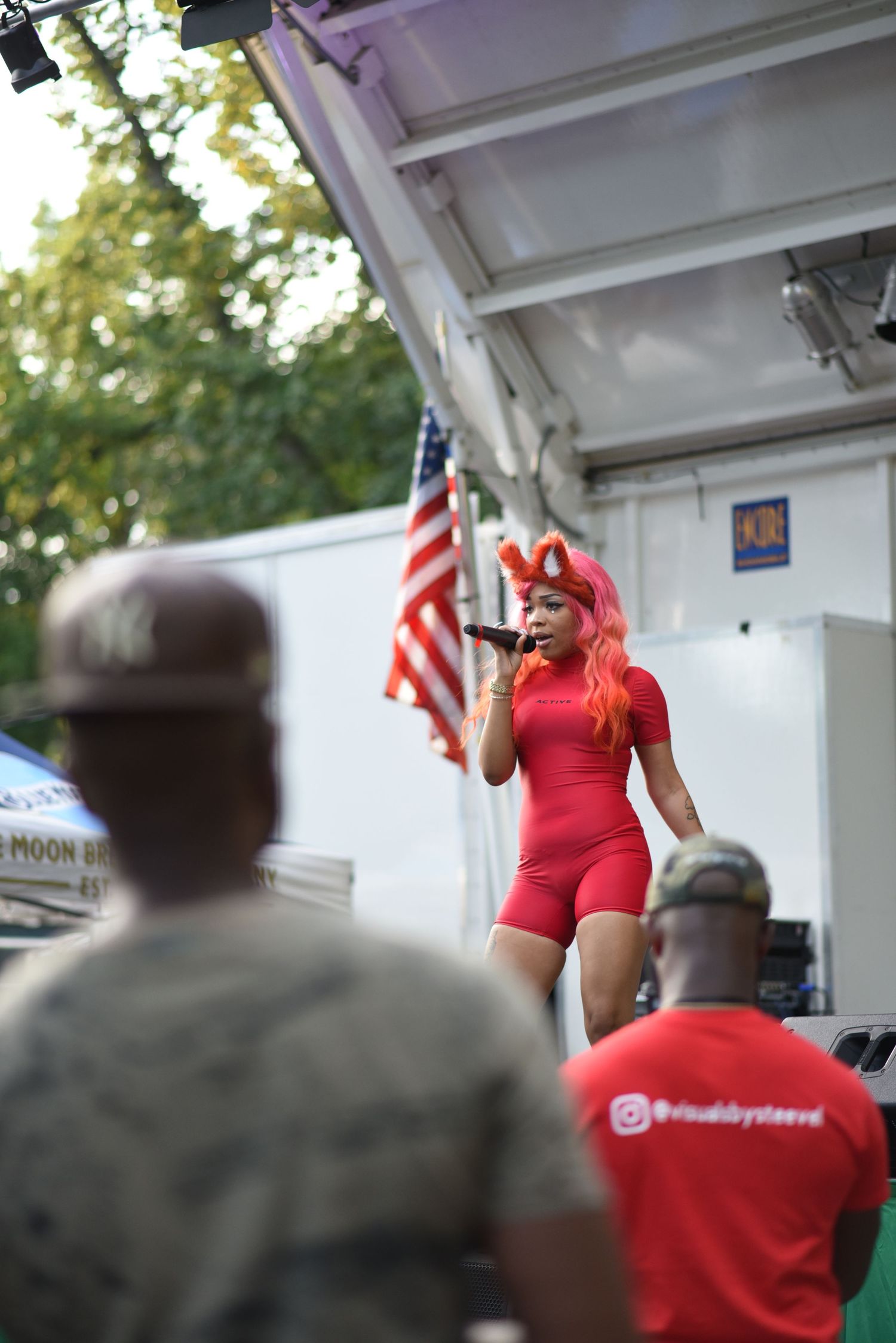 A female performer in red outfit and pink hair sings on outdoor stage with American flag backdrop and audience watching.