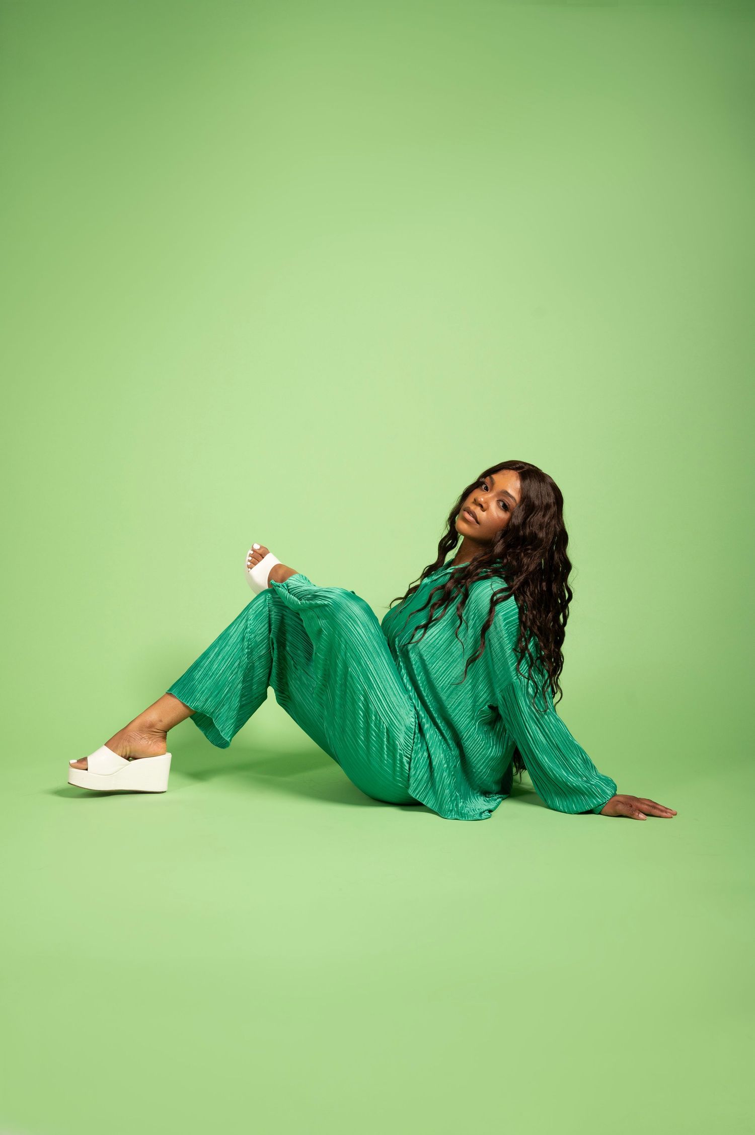 Woman in green outfit sitting on green background wearing white platform sandals.