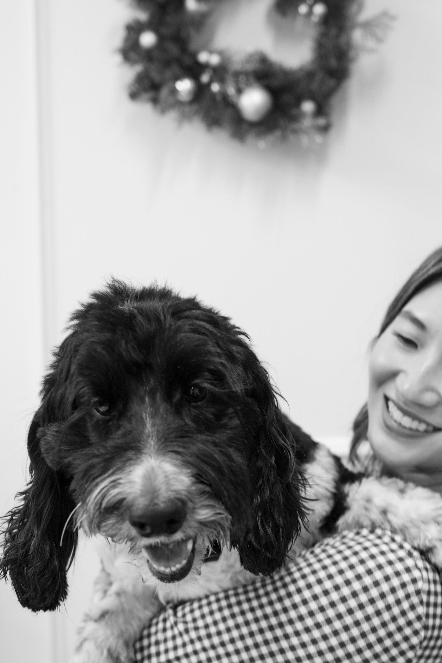 Black and white photo of a smiling woman holding a fluffy black and white dog near a Christmas wreath.