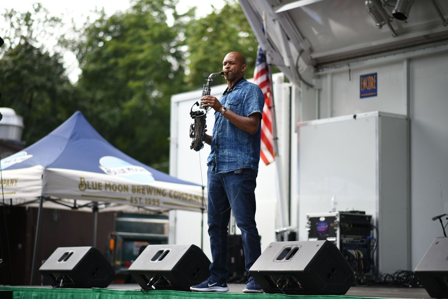 Musician playing saxophone on outdoor stage with American flag and Blue Moon Brewing tent in background.
