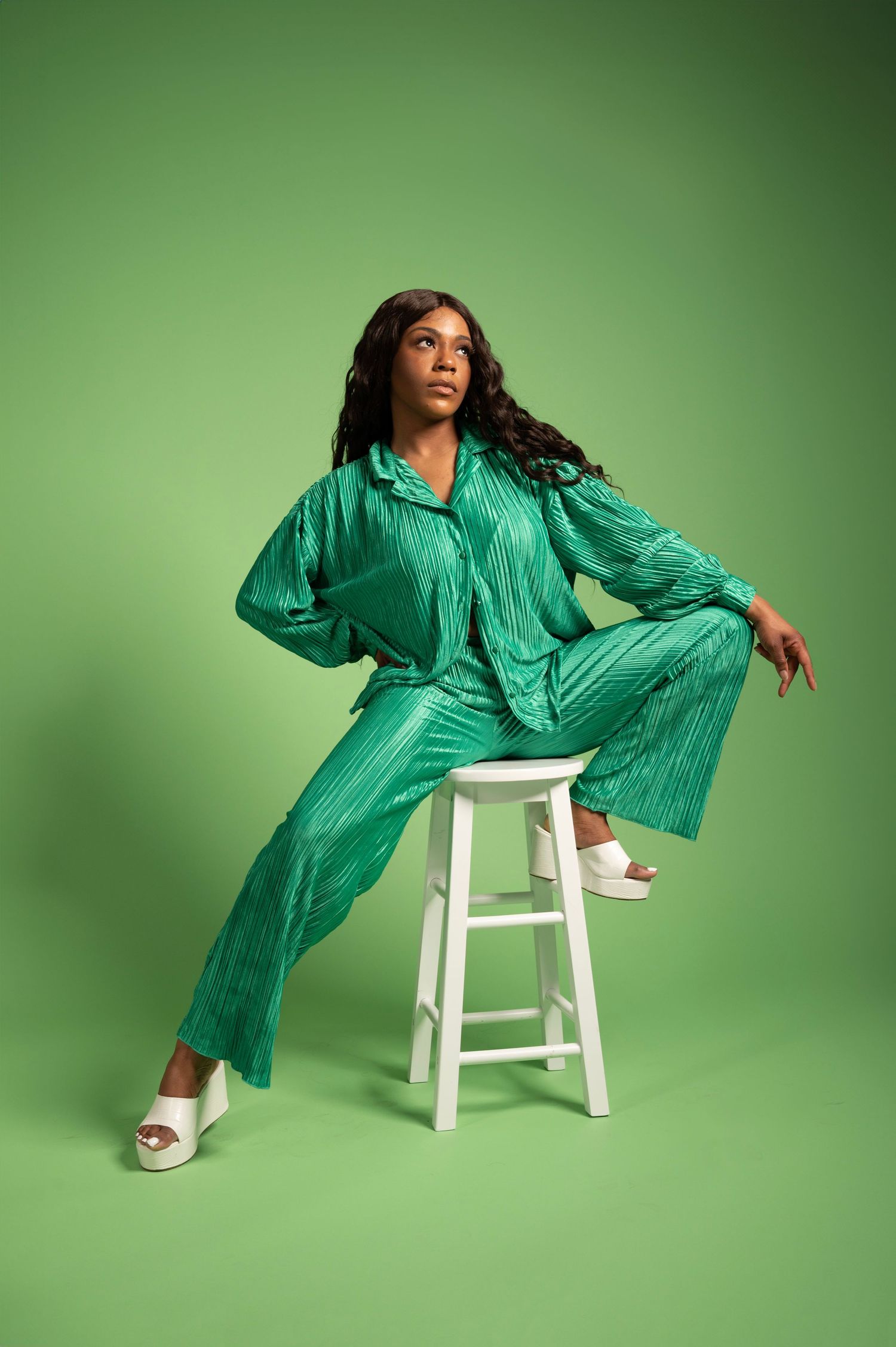 Woman in green plisse co-ord set seated on white stool against green background, wearing white heeled sandals.