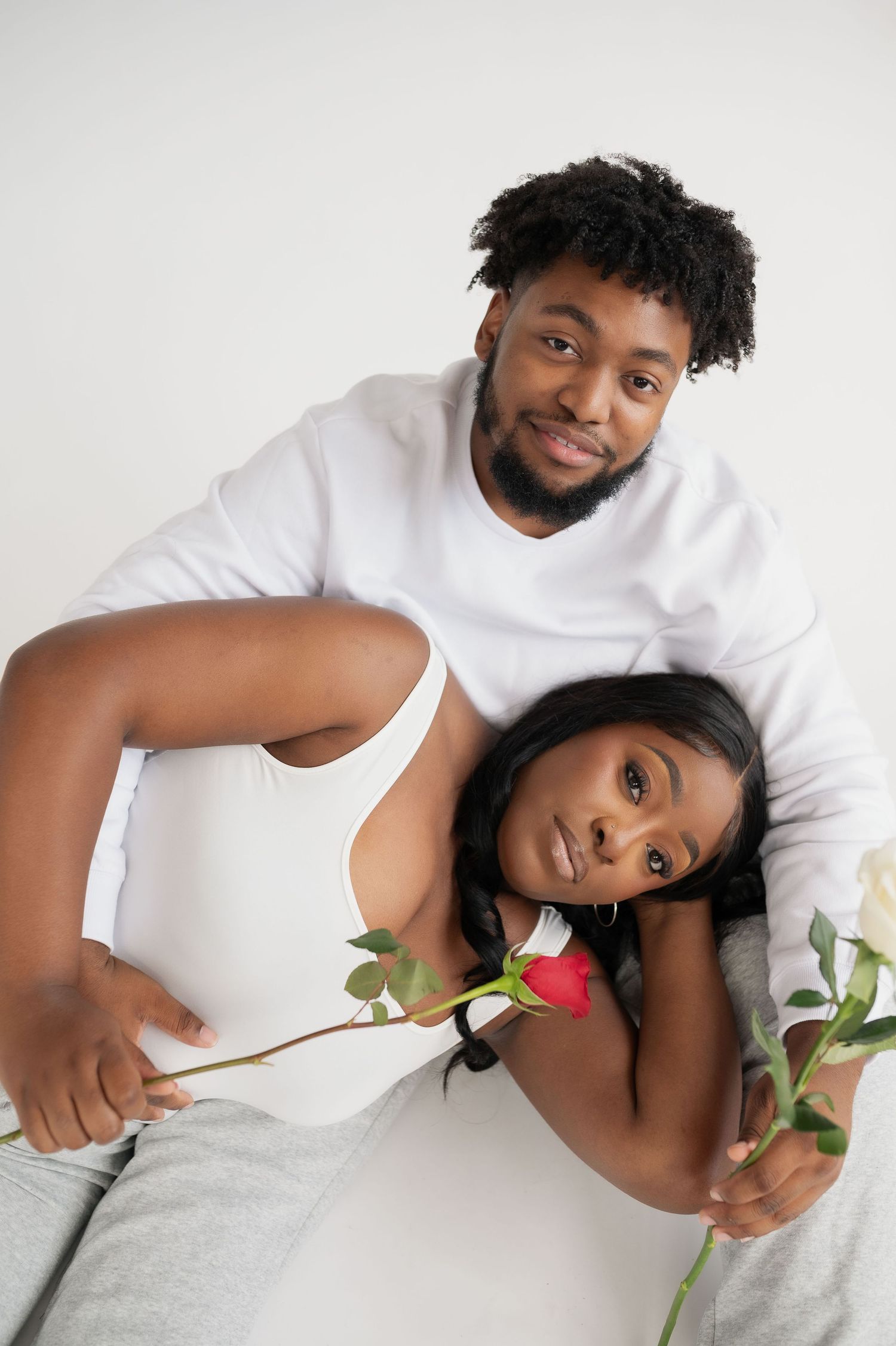 A romantic couple in white outfits lying down with roses in a studio setting.