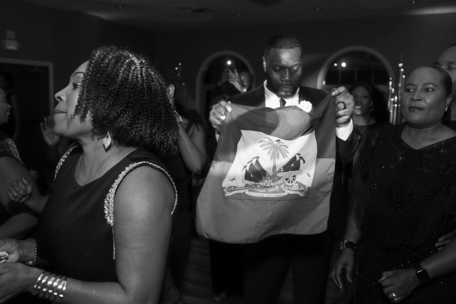Black and white photo of people dancing at a party, man holding a Haitian flag in the center.