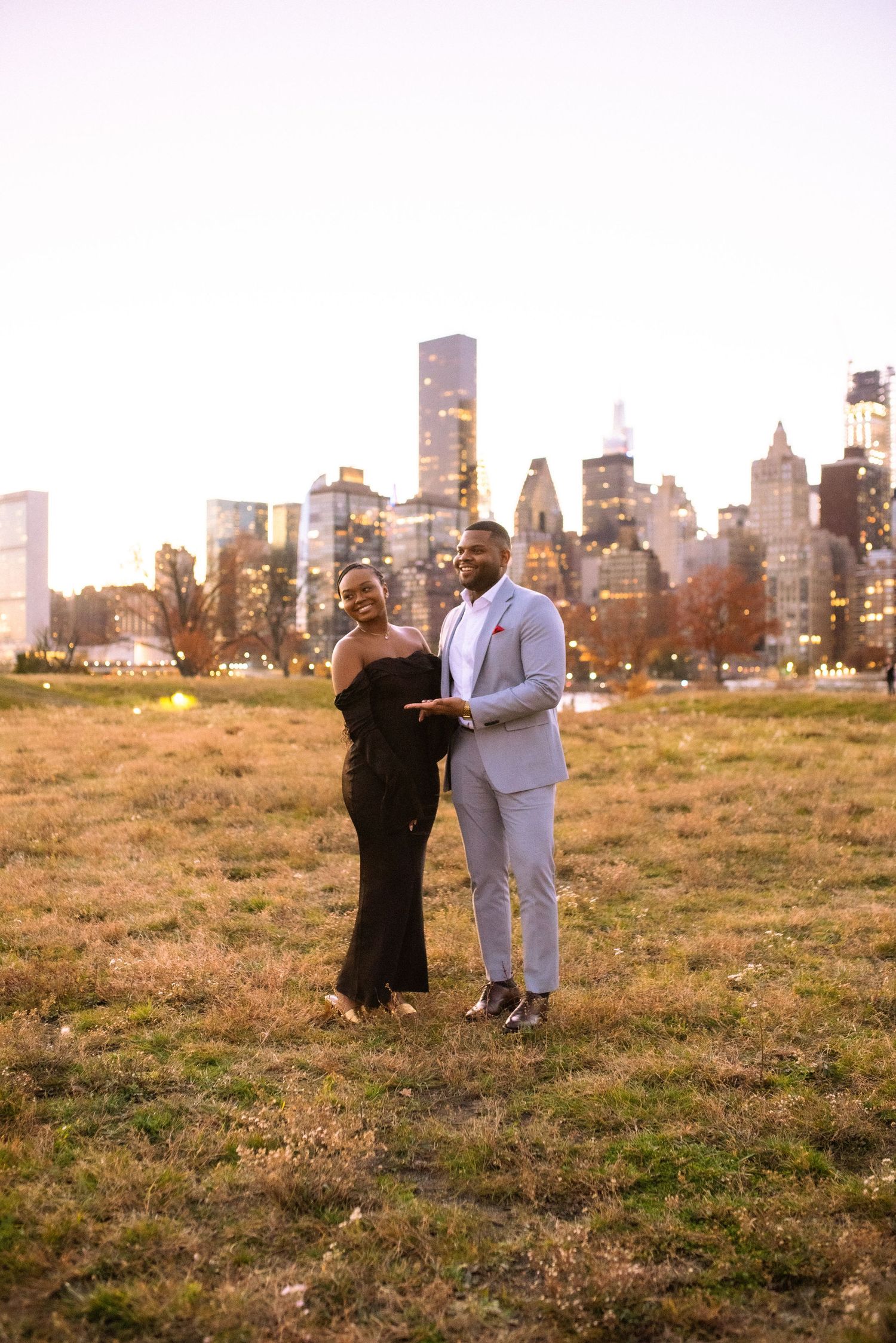 Two people in contrasting outfits standing in a field with New York City skyline at sunset.