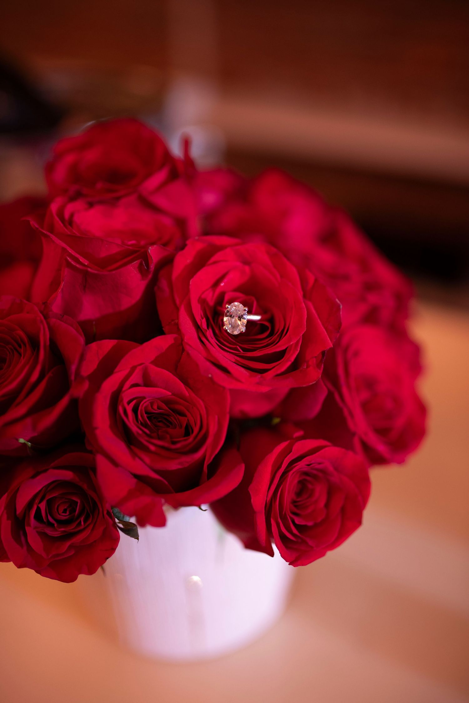 Deep red roses arranged in a white vase with a sparkling diamond ring nestled among the petals.