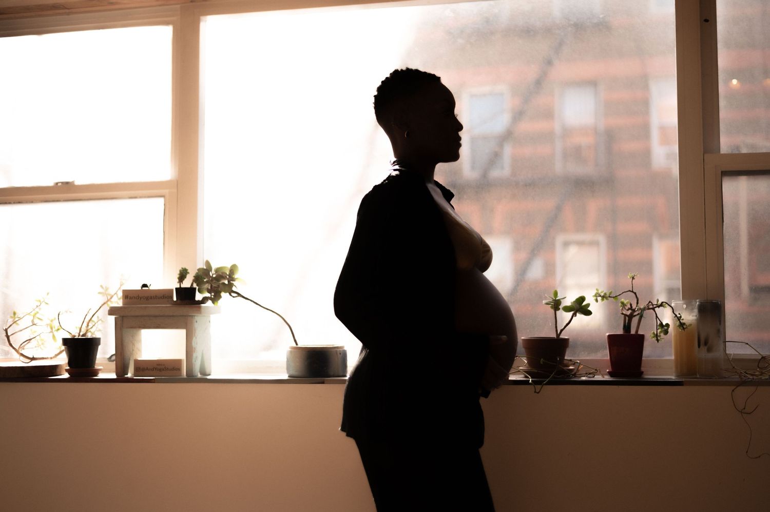 Silhouetted figure by window during golden hour with urban buildings visible outside holding her baby bump.