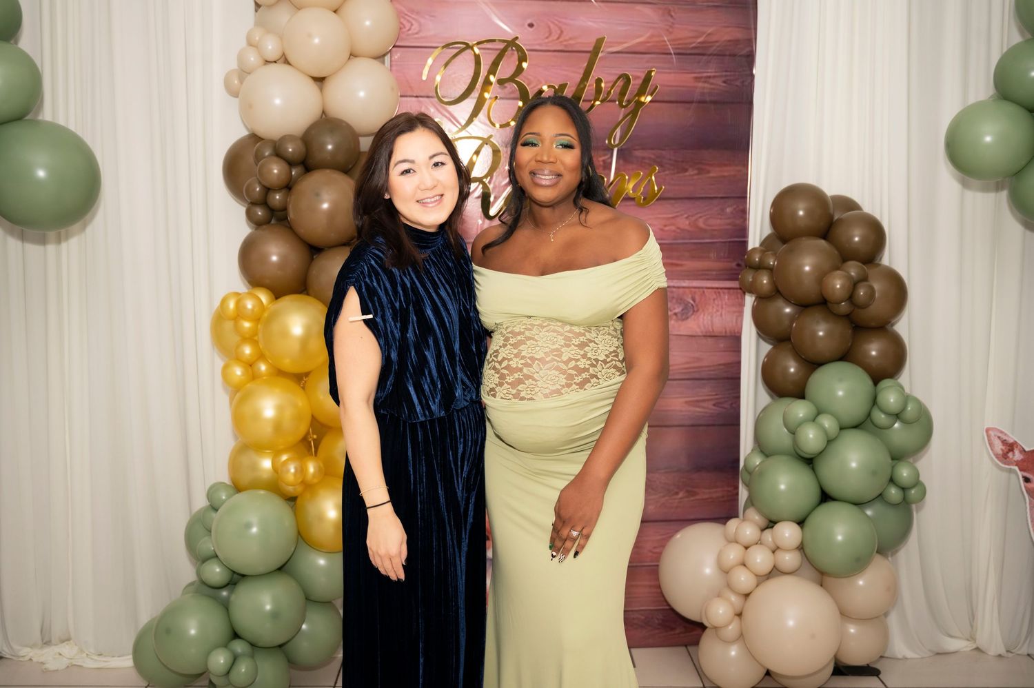Two women posing together at a baby shower in front of a balloon arch backdrop with sage green and brown balloons.