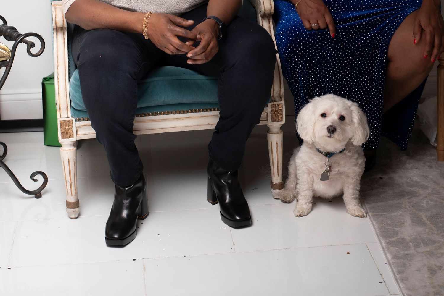 Two people sitting on ornate chairs with a small white fluffy dog standing beside them on a tiled floor.