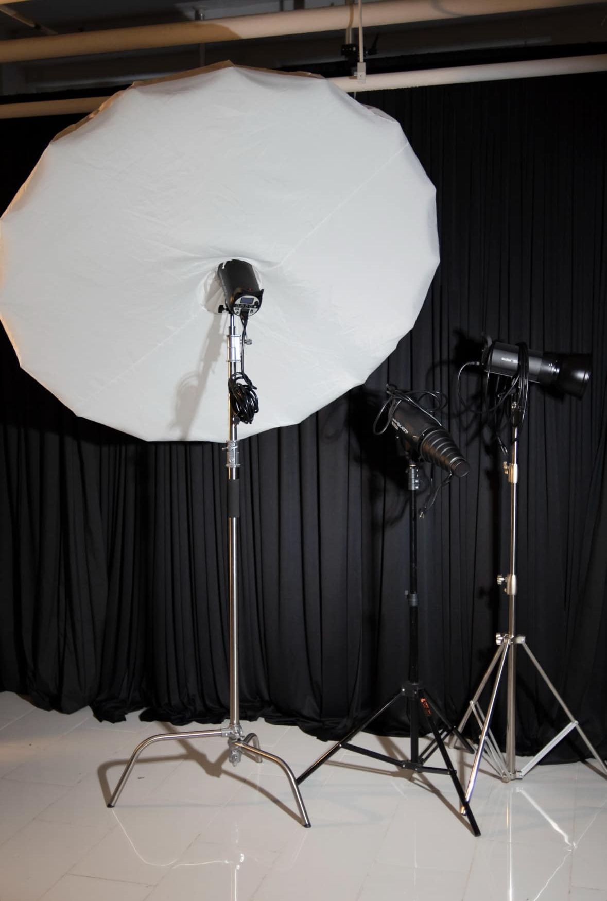 Professional photography studio setup with large white umbrella light, strobe lights on stands, and black backdrop curtain.