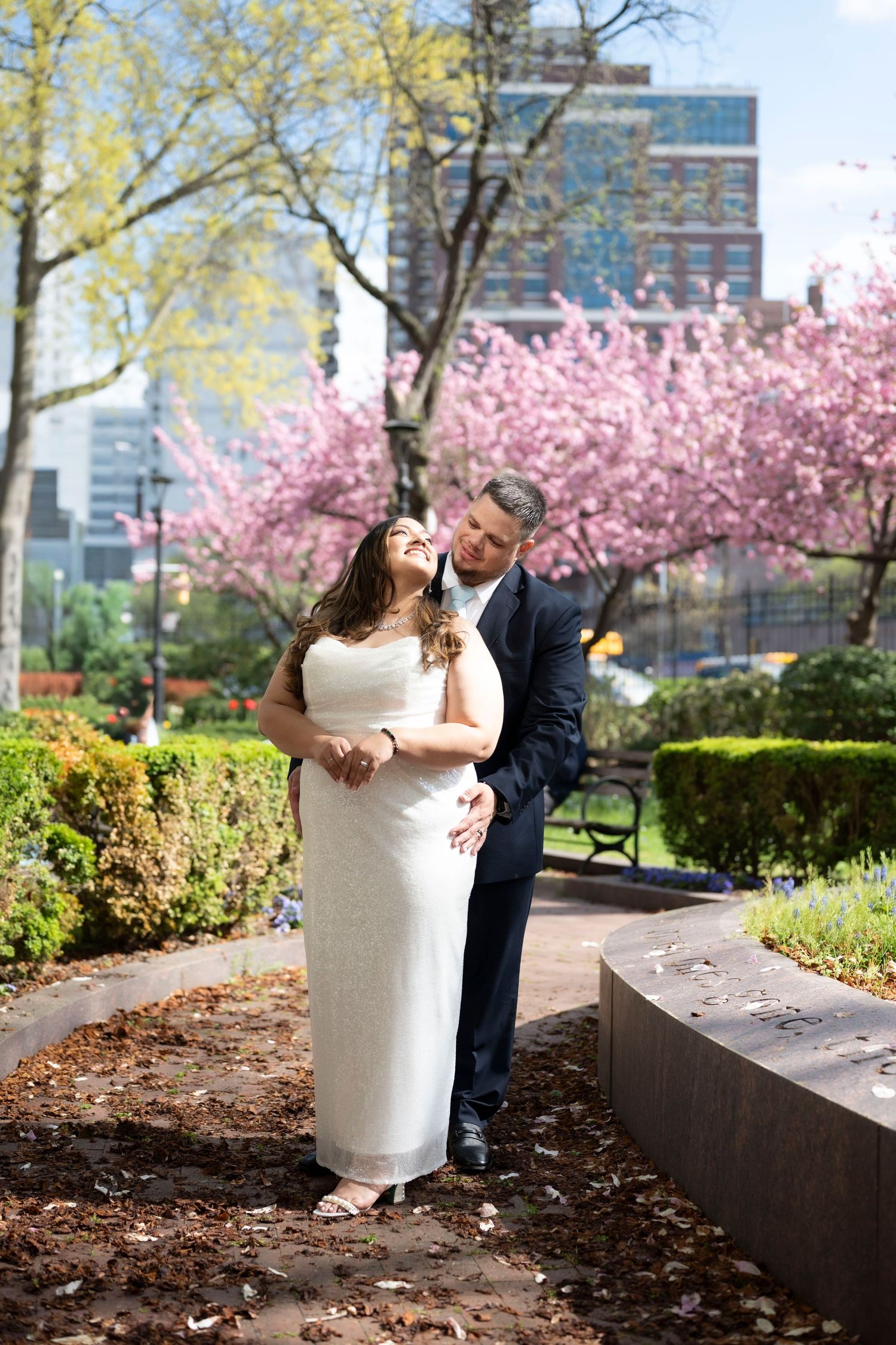 Couple embracing in a park with pink cherry blossoms and city buildings in the background on a sunny spring day.