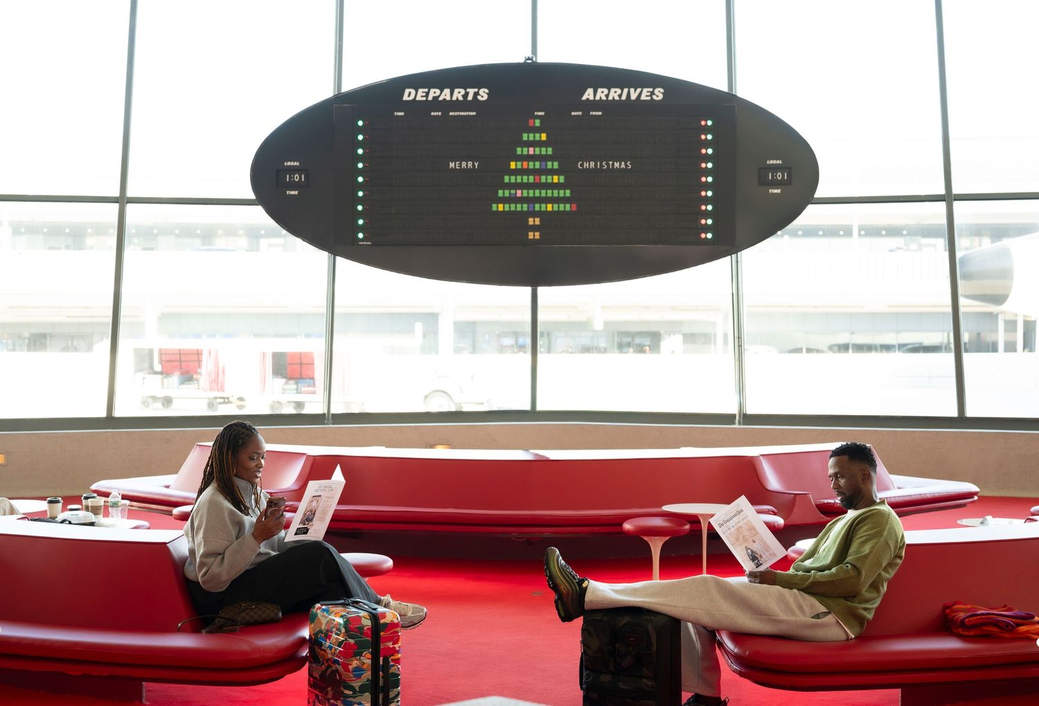 Two people relaxing in modern red lounge area beneath digital display board.