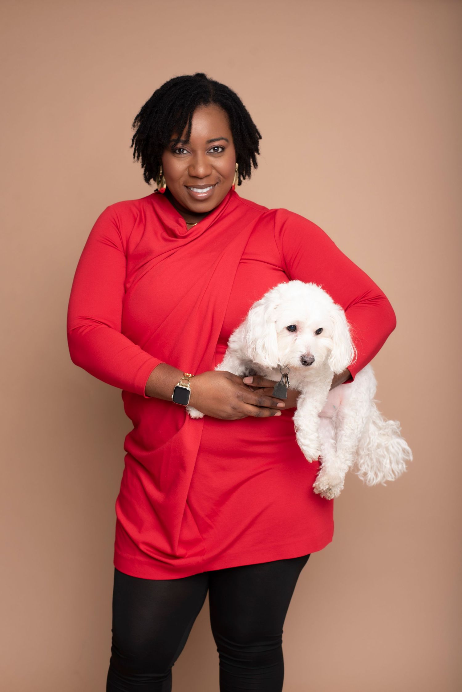 Woman in red turtleneck holding a fluffy white Bichon Frise dog against a tan studio background.