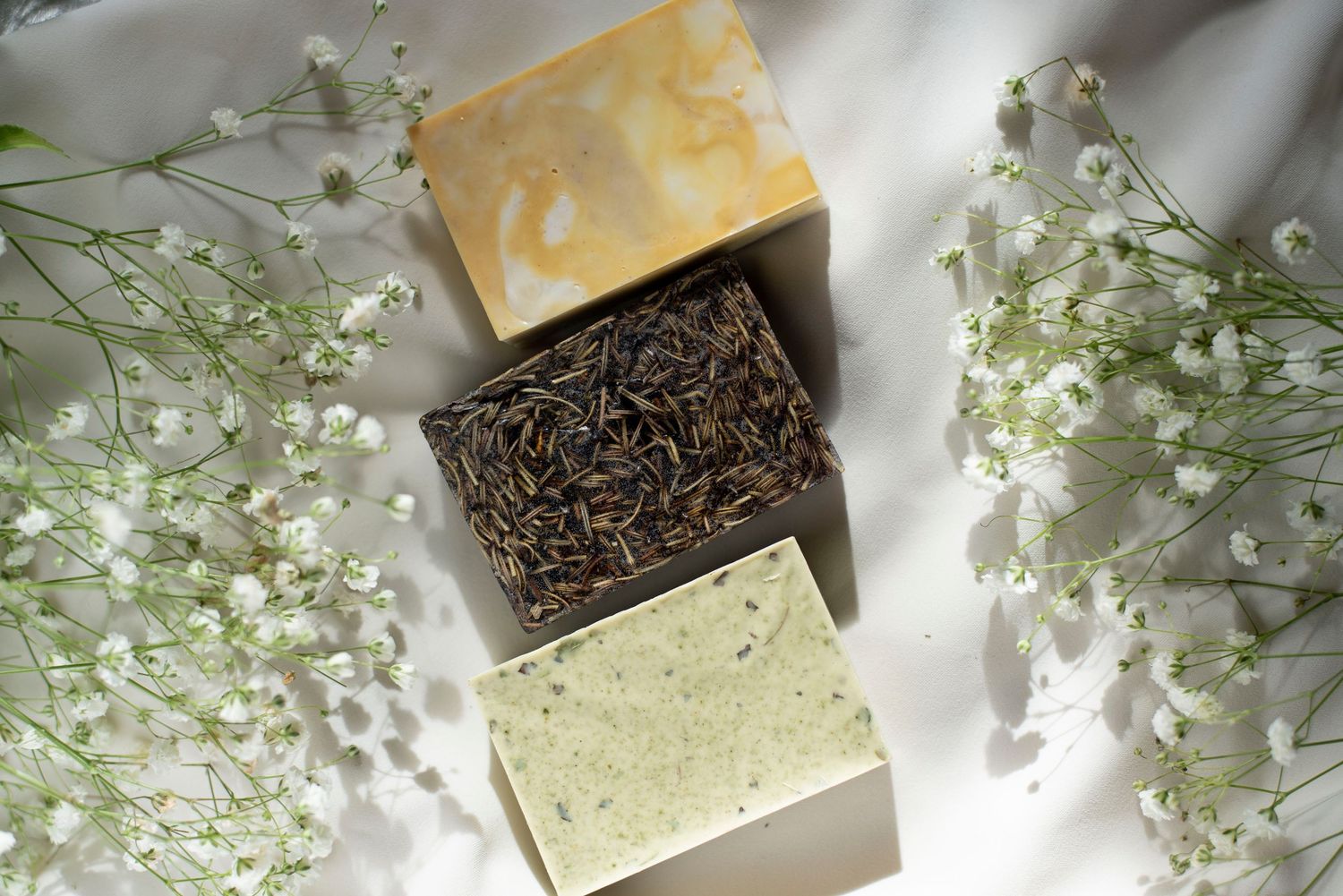 Three artisanal soap bars with baby's breath flowers on white fabric background.