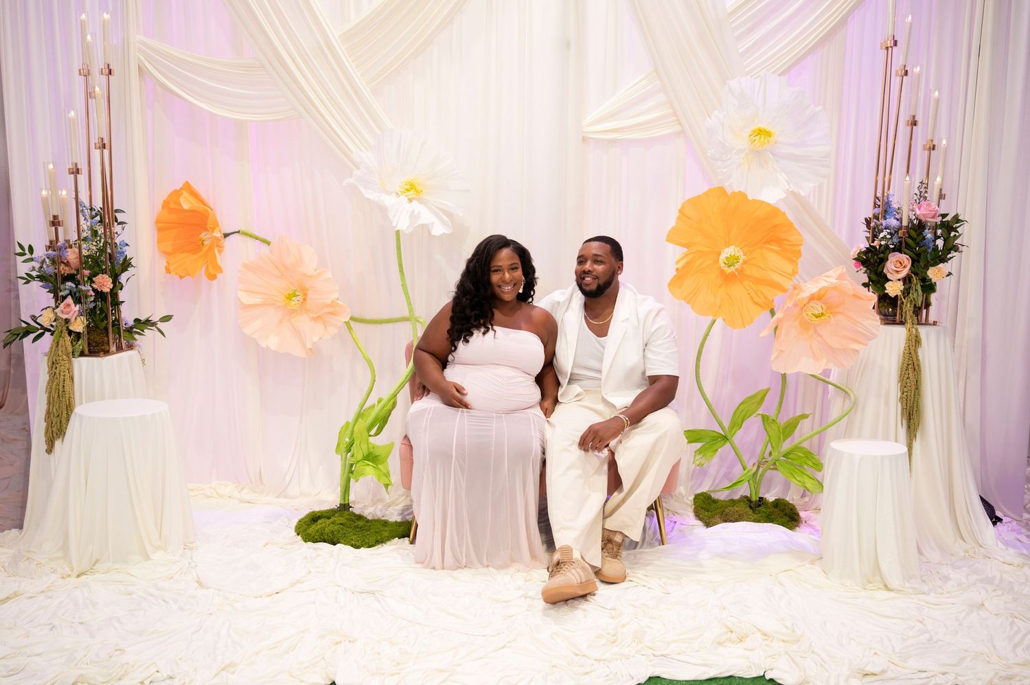 Elegant white draped photo set with large floral decorations in soft lighting. Couple sitting in the middle