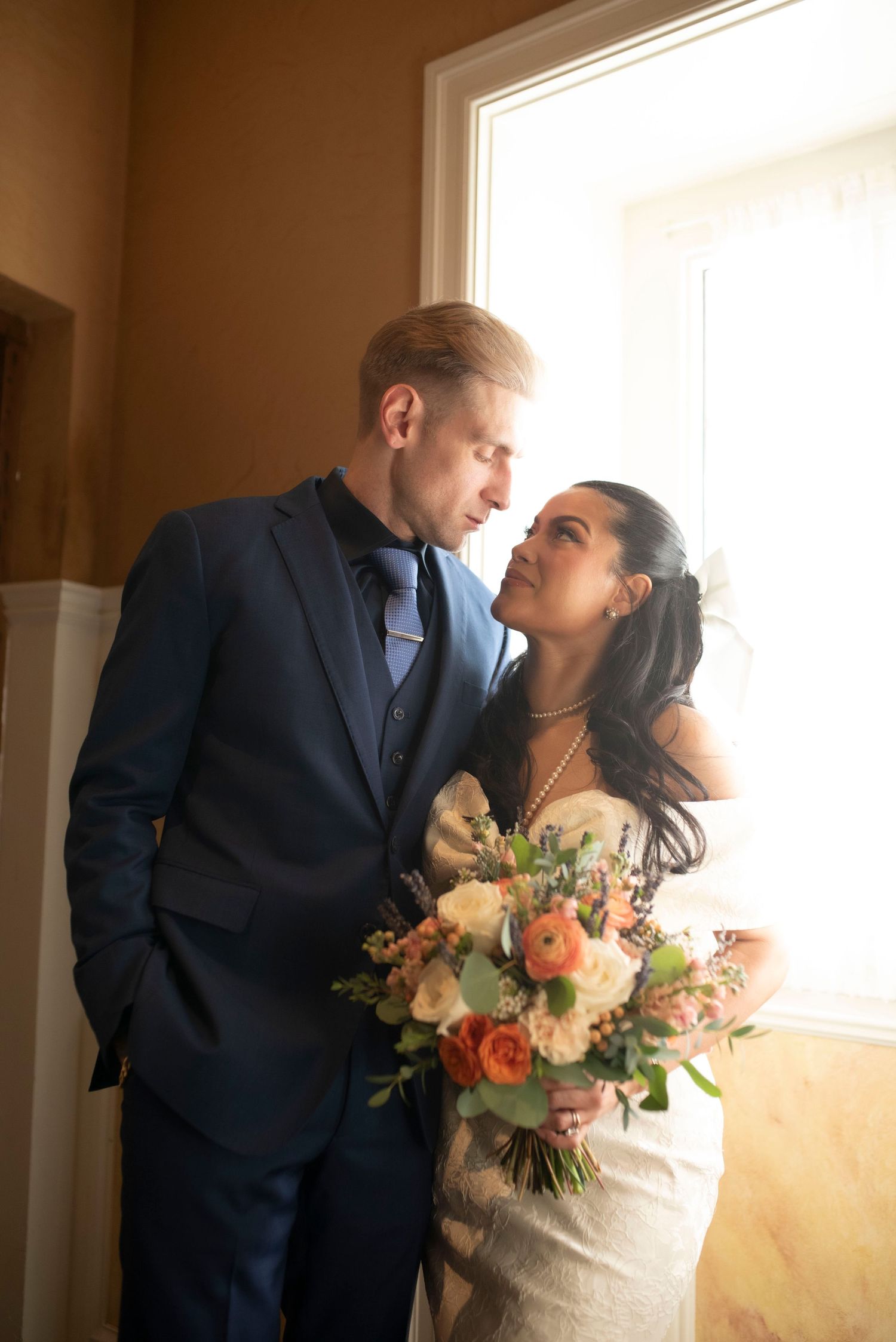 Wedding couple shares intimate moments near window with beautiful floral bouquet.